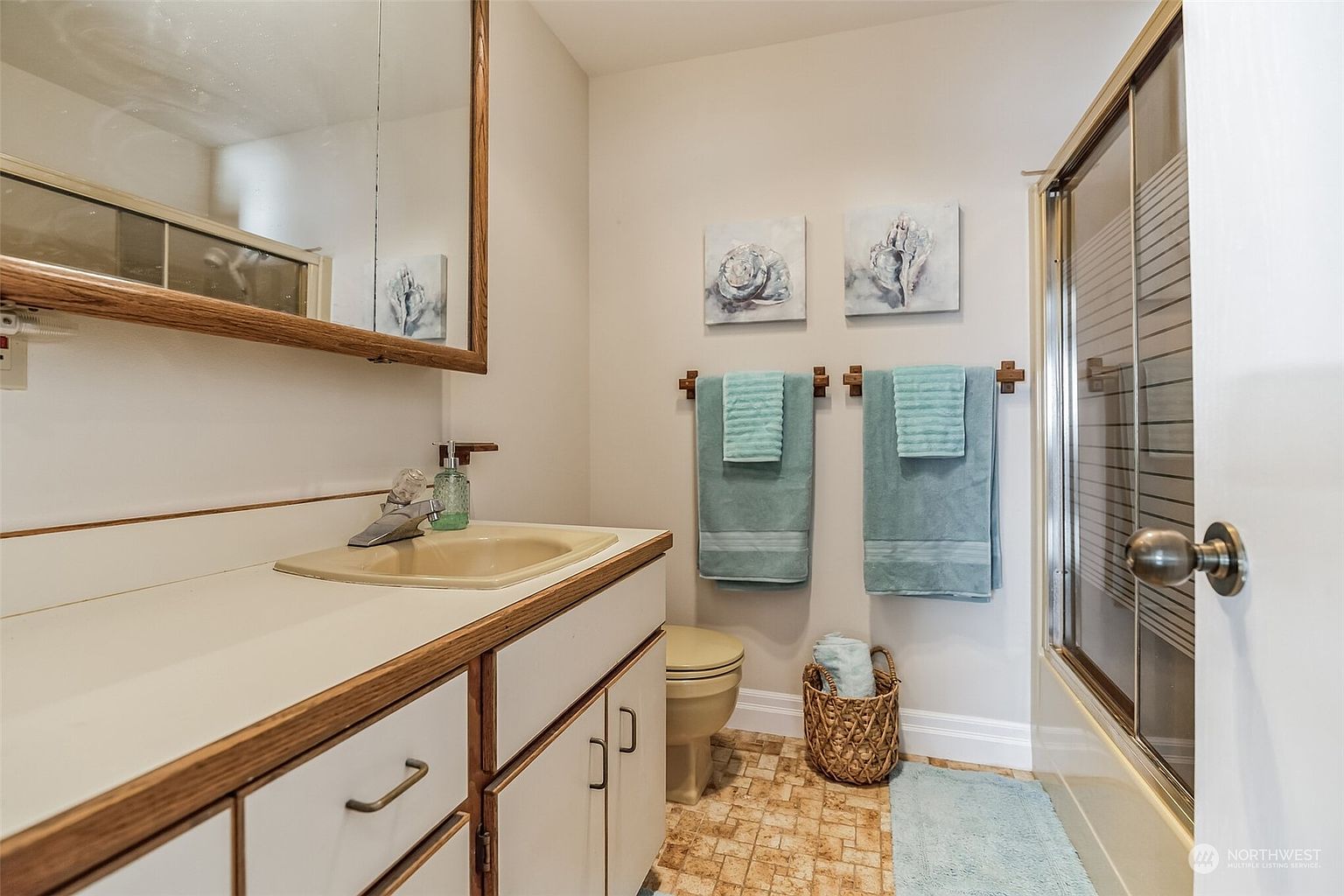 This functional bathroom features a vanity with a light-colored sink, a wood-framed mirror, and a shower stall with sliding glass doors. The walls are decorated with two seashell-themed art pieces above a towel rack holding teal towels, while a woven basket sits on the patterned tile floor. The space is compact and brightly lit, offering a clean and simple aesthetic.