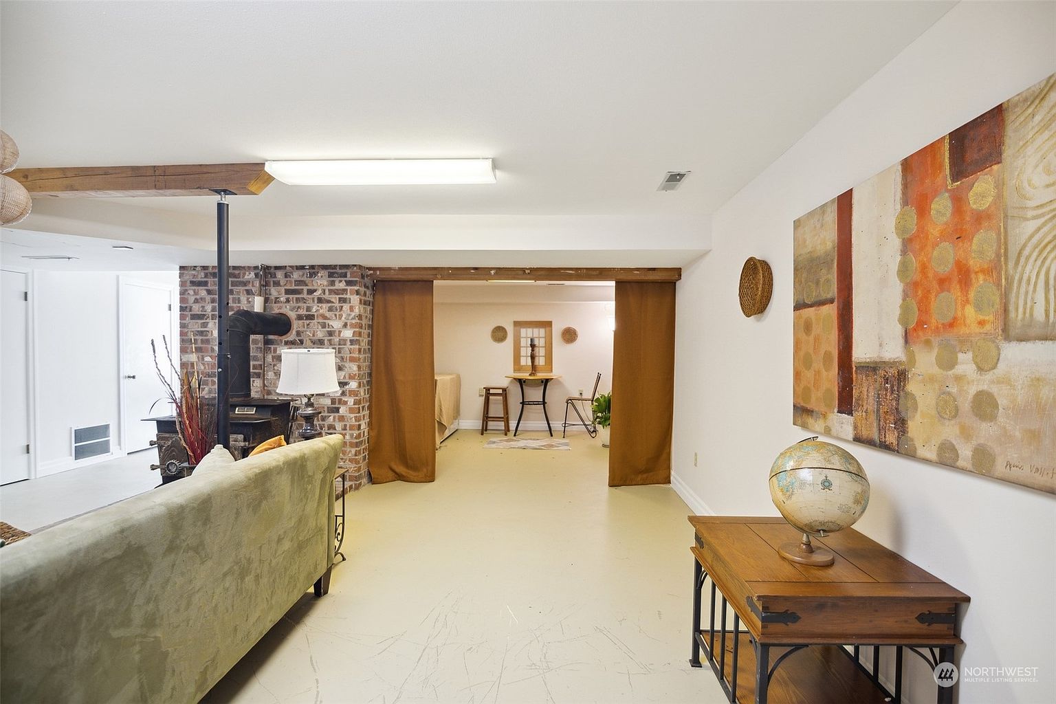 This basement living area features a neutral color palette with a brick chimney breast housing a wood-burning stove. A green sofa sits in the foreground, while a pair of brown curtains separates the main space from a secondary area containing a small bistro table and chairs. The room is illuminated by a long, rectangular overhead light fixture and decorated with a large abstract wall painting and a globe on a wooden console table.