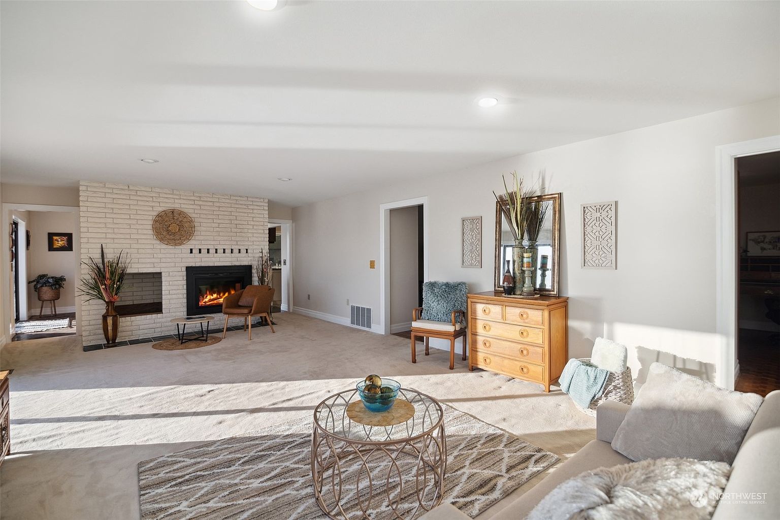 This spacious living room features a light-toned carpet and a prominent white-painted brick fireplace as a central focal point. The room is furnished with a comfortable sofa, a round metal-framed coffee table, and a wooden chest of drawers topped with a decorative mirror. The perspective captures the open-concept layout, highlighting the neutral color palette and the natural light streaming into the space.