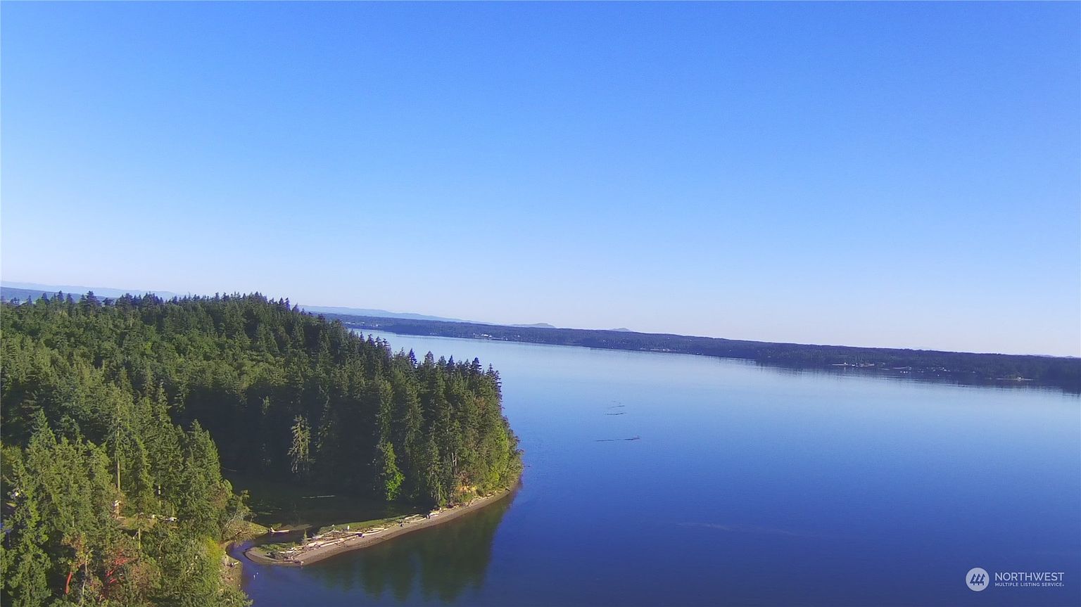 This high-angle aerial view captures a serene coastal landscape featuring a dense, lush forest meeting a calm, expansive body of water. A small, sandy beach curves along the shoreline, providing a natural transition between the wooded terrain and the deep blue water. The perspective emphasizes the vastness of the waterfront setting, highlighting the property's secluded and tranquil location.