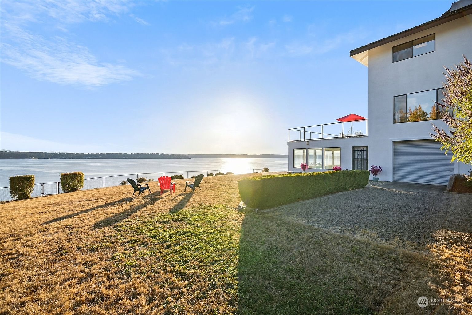 This rear view captures a multi-level white home situated on a waterfront property, featuring a spacious gravel driveway and a large, sun-drenched lawn. Three chairs are positioned on the grass, overlooking a vast body of water under a bright, clear sky. The home includes a second-story balcony with a red umbrella, large windows, and a garage, offering a serene and expansive outdoor living space.