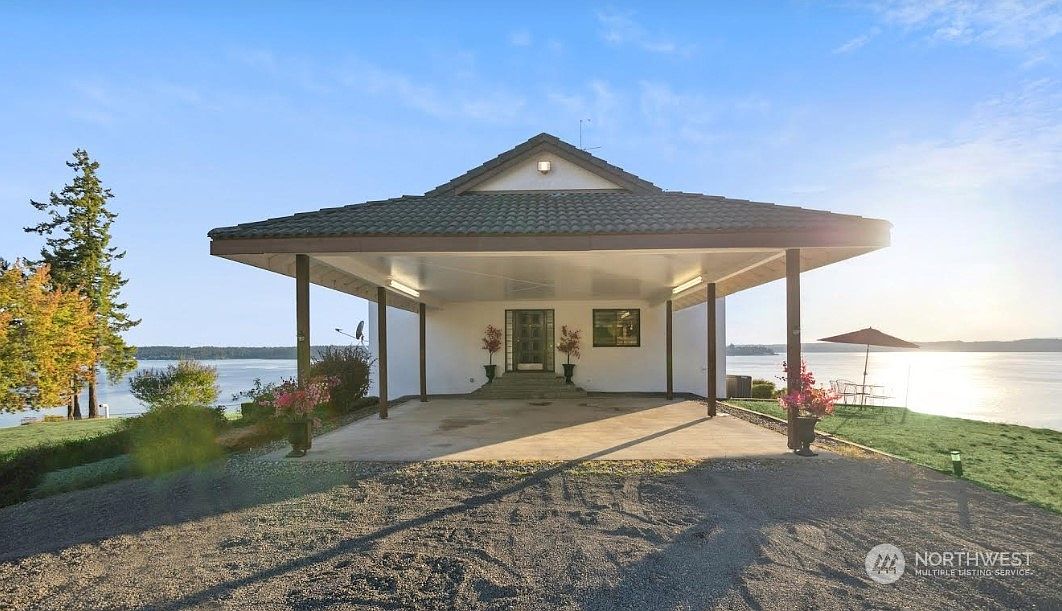 This front-facing view captures a unique home entrance featuring a large, covered carport area that leads directly to the front door. The structure is set against a scenic waterfront backdrop, with a gravel driveway leading up to the concrete pad. The home's design is simple and functional, emphasizing the expansive views of the water and the surrounding landscape.
