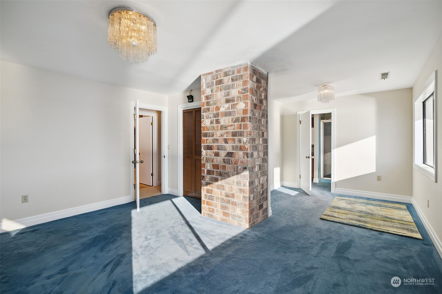 This bedroom features blue carpeting and a central brick chimney breast that divides the space. The room is illuminated by two crystal-style flush-mount light fixtures and receives natural light from a window on the right. Two doorways lead to adjacent rooms, and a small area rug sits on the floor, creating a distinct layout with a vintage aesthetic.