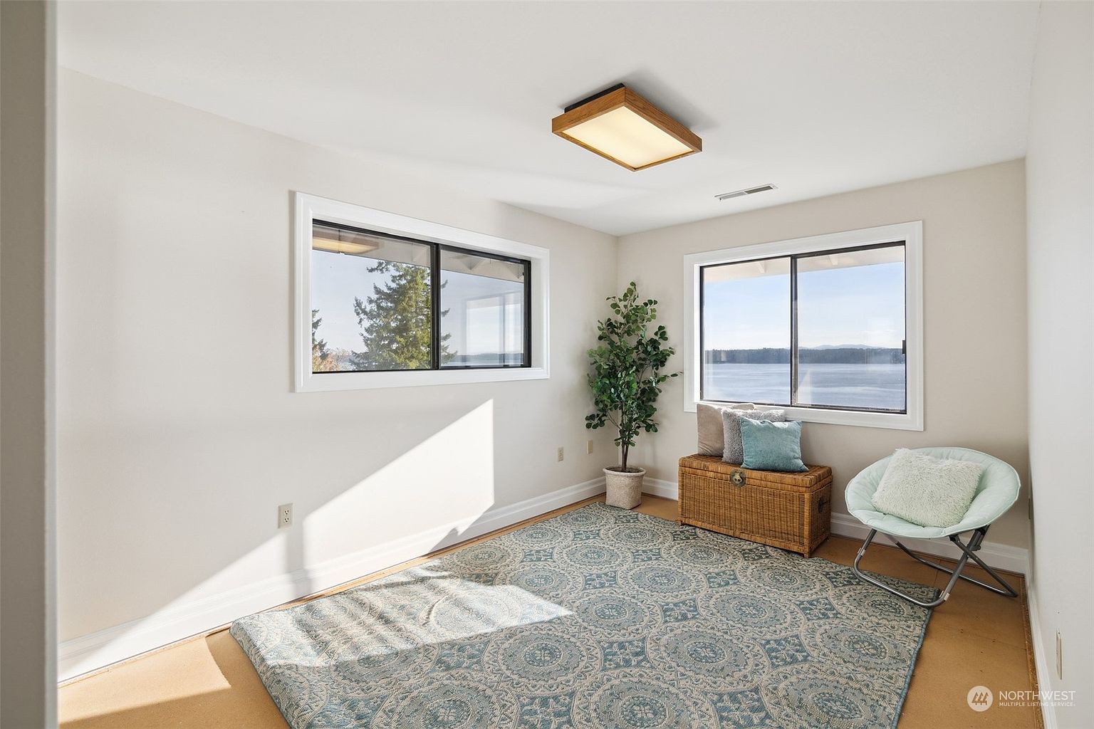 This bright, neutral-toned room features large windows that offer expansive views of the water, creating a serene and airy atmosphere. The space is furnished with a woven storage chest topped with decorative pillows and a modern, light-colored saucer chair, all set upon a large, patterned area rug. A potted faux tree adds a touch of greenery to the corner, while a simple wooden flush-mount light fixture provides overhead illumination.