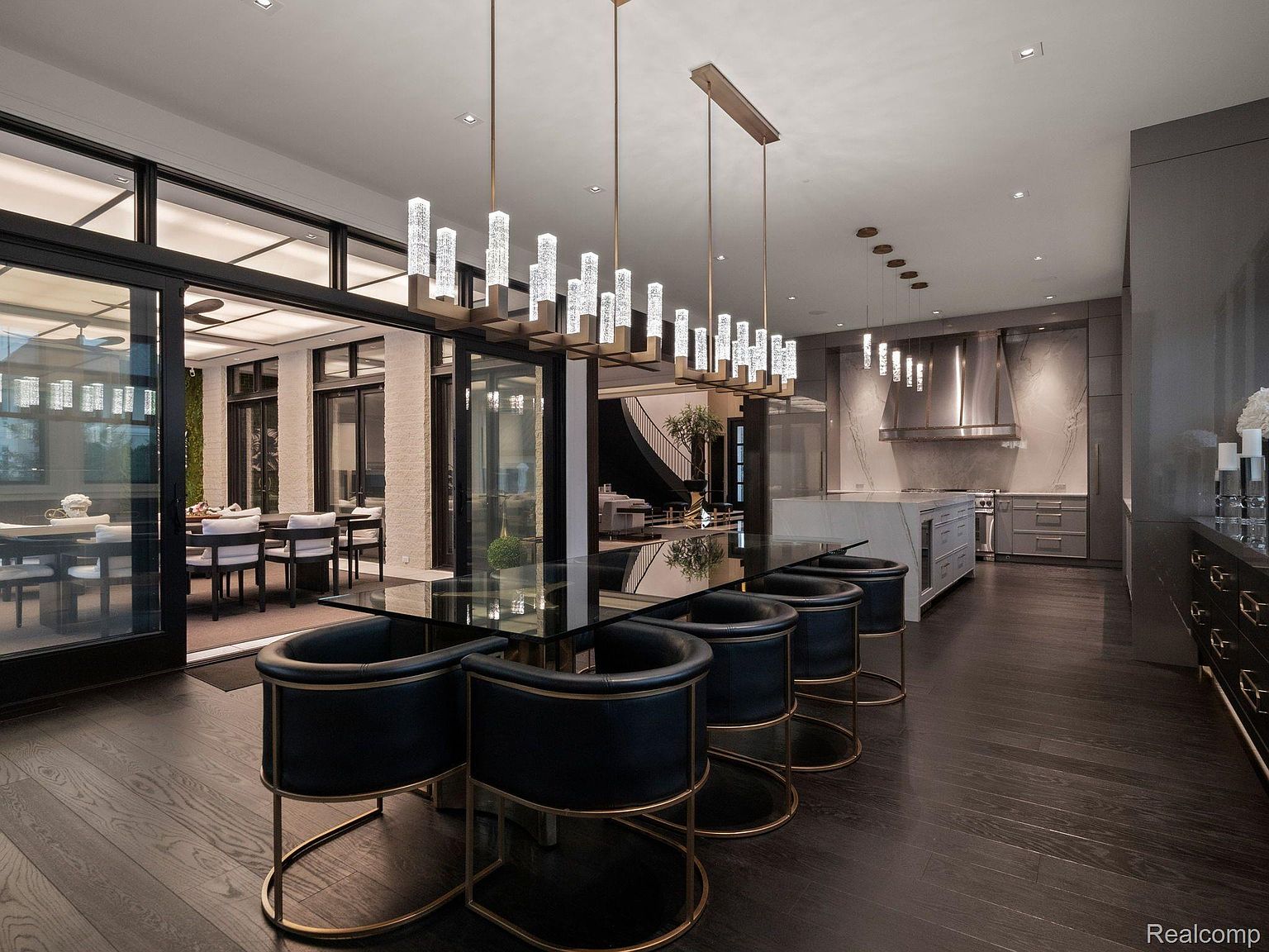 This is an interior shot of a modern dining room featuring a long, black glass dining table with black and gold chairs. Above the table hangs a contemporary chandelier with crystal accents. The room opens to an outdoor dining area through large glass doors, and a modern kitchen is visible in the background, creating a seamless transition between spaces.