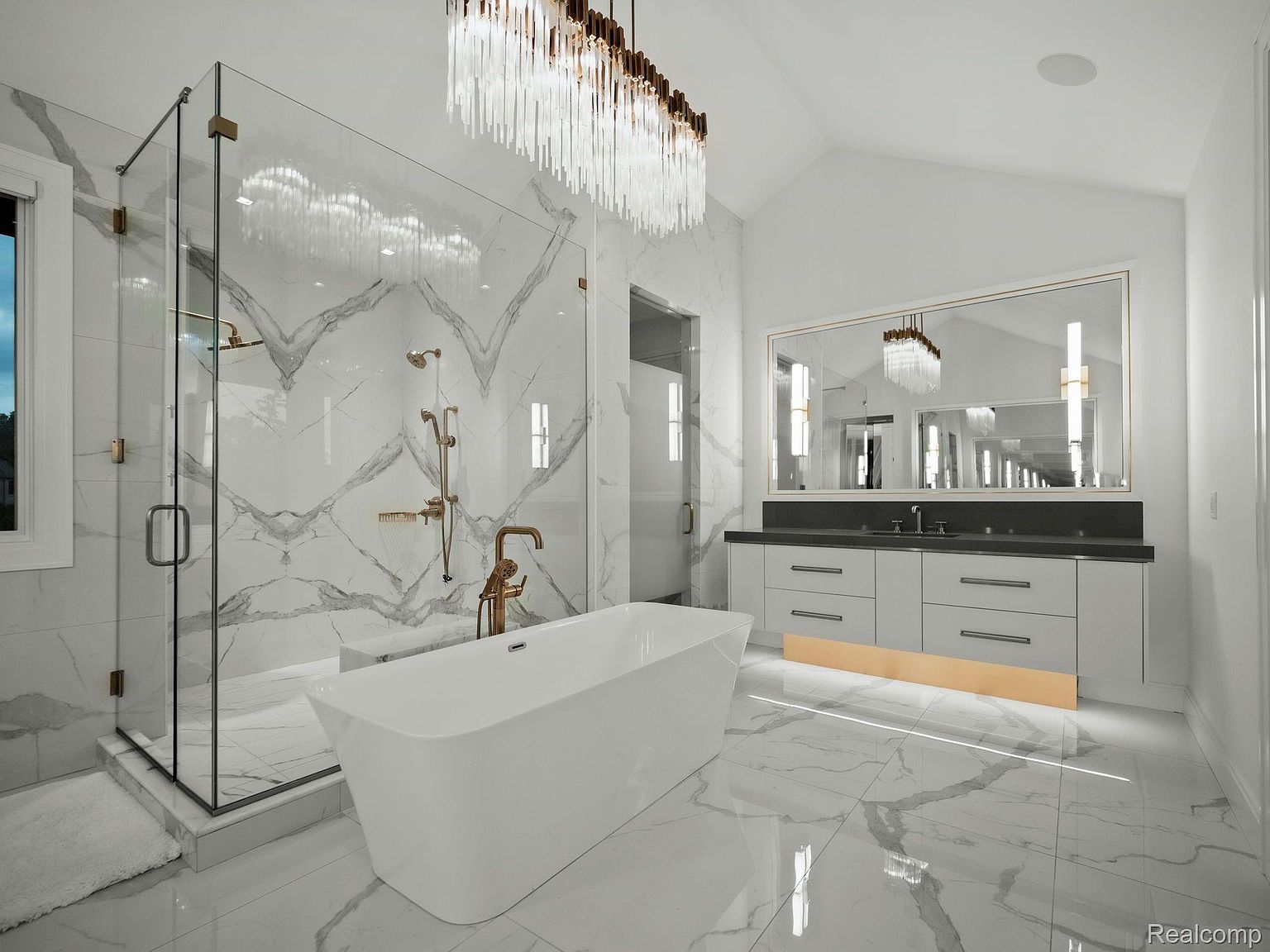 This is a luxurious primary bathroom featuring a freestanding white bathtub, a glass-enclosed shower with gold fixtures, and a double vanity with a large mirror. The walls and floors are covered in marble with gray veining, and a modern chandelier hangs from the ceiling. The overall impression is one of elegance and sophistication.
