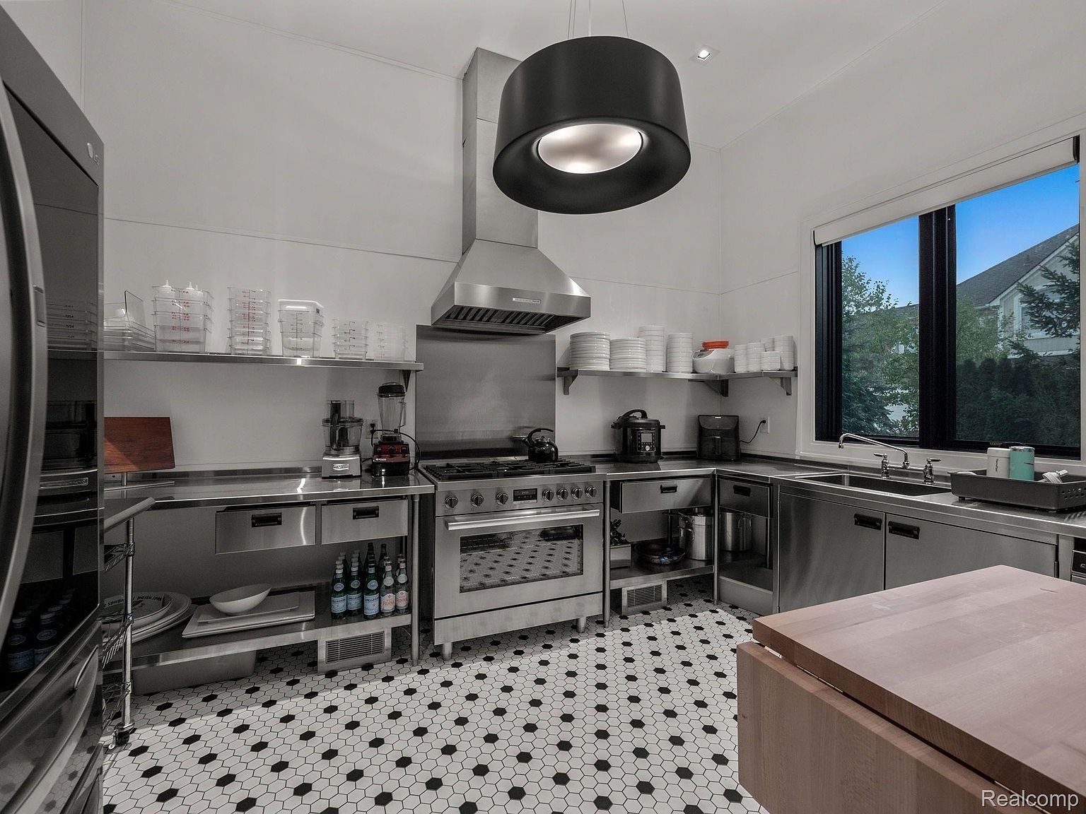This is a commercial-style kitchen featuring stainless steel countertops and cabinetry, a professional-grade range with a hood, and open shelving. The black and white hexagonal tile flooring adds a classic touch, while the large window provides natural light and a view of the outdoors. The overall impression is clean, functional, and well-equipped for serious cooking.