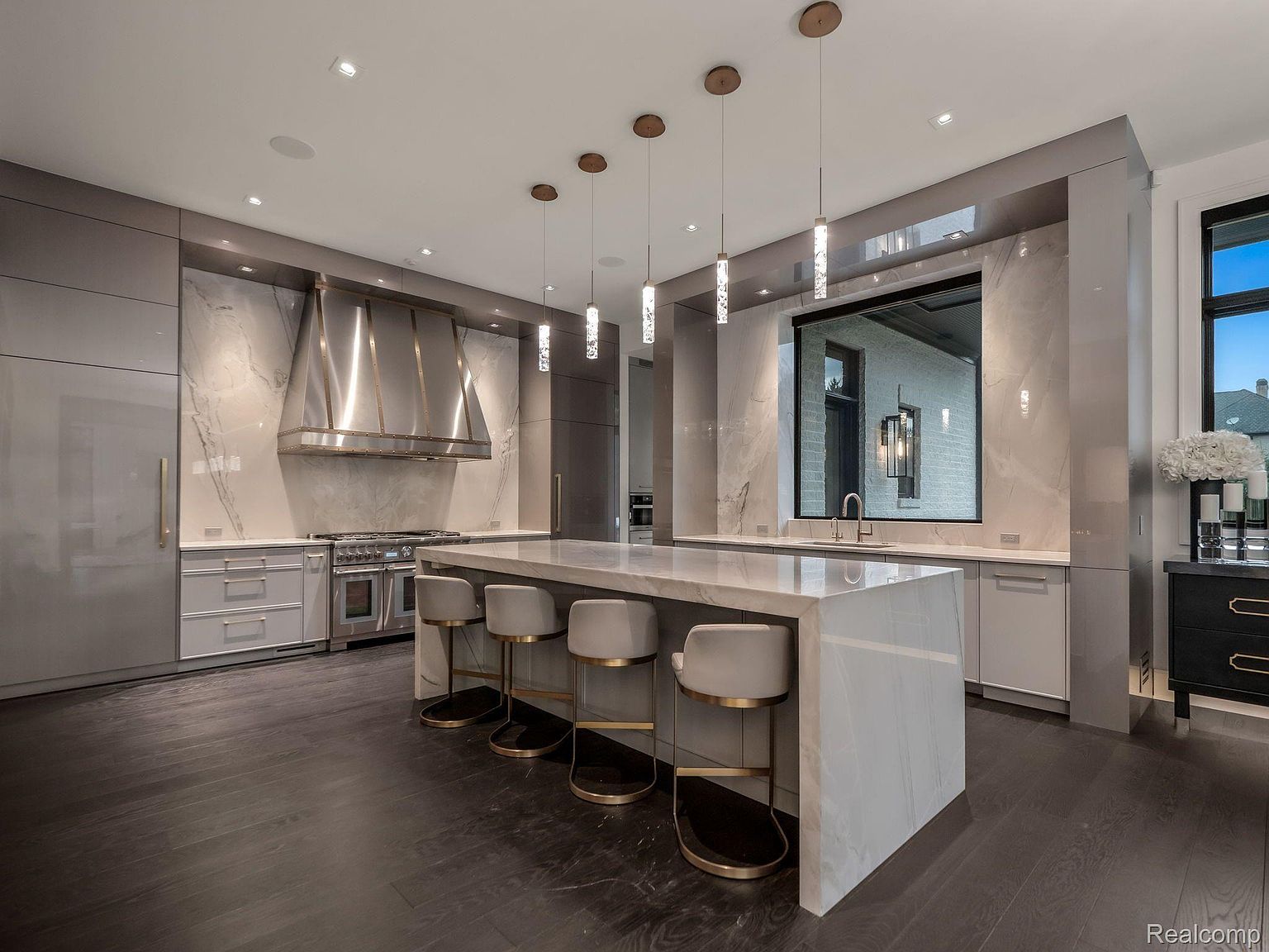 This is a modern, high-end kitchen featuring a large island with seating, marble countertops and backsplash, and stainless steel appliances. The kitchen is well-lit with pendant lighting and recessed lighting, and the dark wood floors provide a nice contrast to the light-colored cabinetry and marble. The perspective is from a wide angle, showcasing the entire kitchen space.