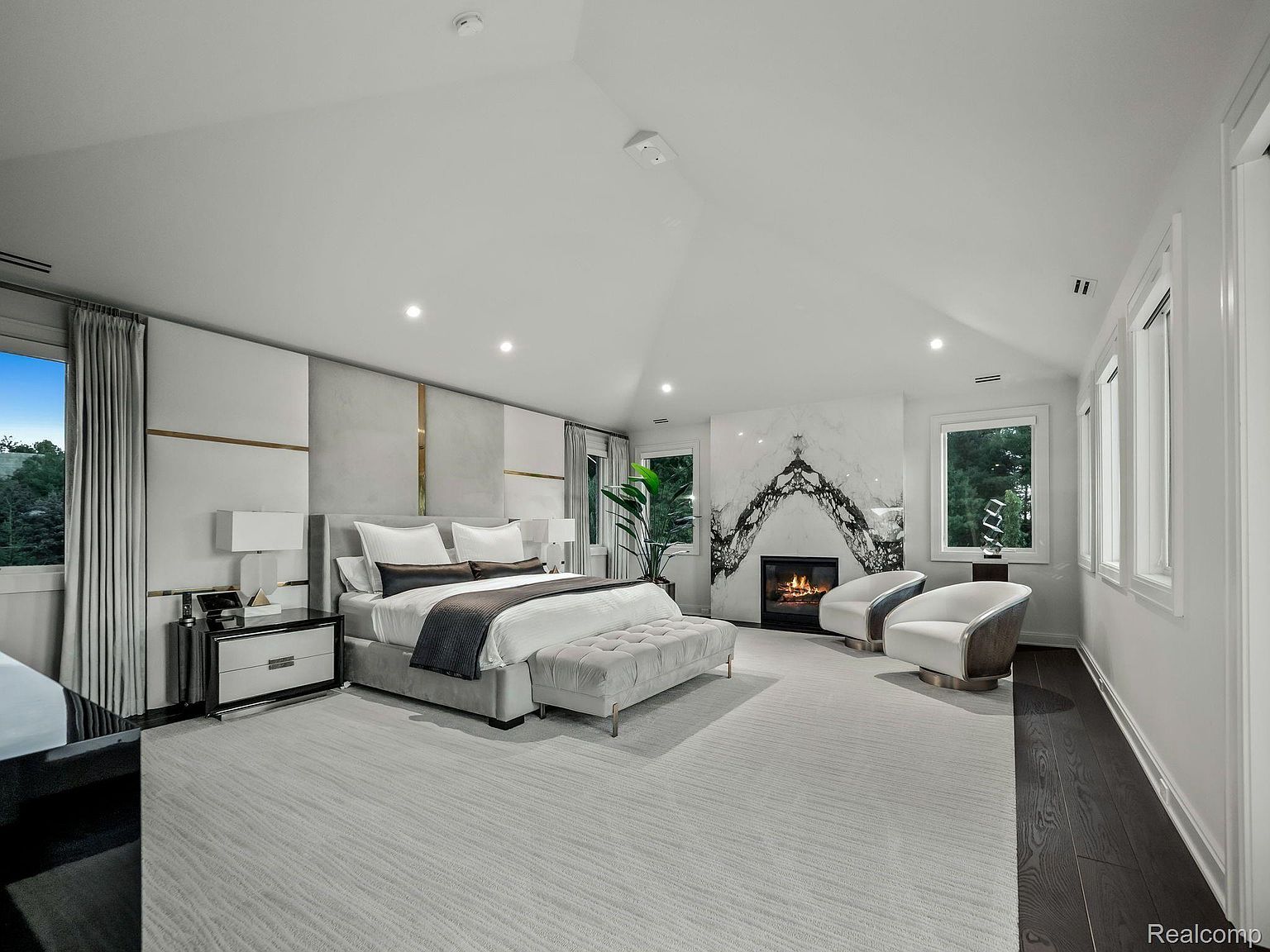 This is a spacious primary bedroom featuring a king-size bed with a gray upholstered frame and a marble fireplace. The room is decorated in a modern style with white walls, light gray carpet, and gold accents. Large windows provide natural light, and there are two armchairs near the fireplace.