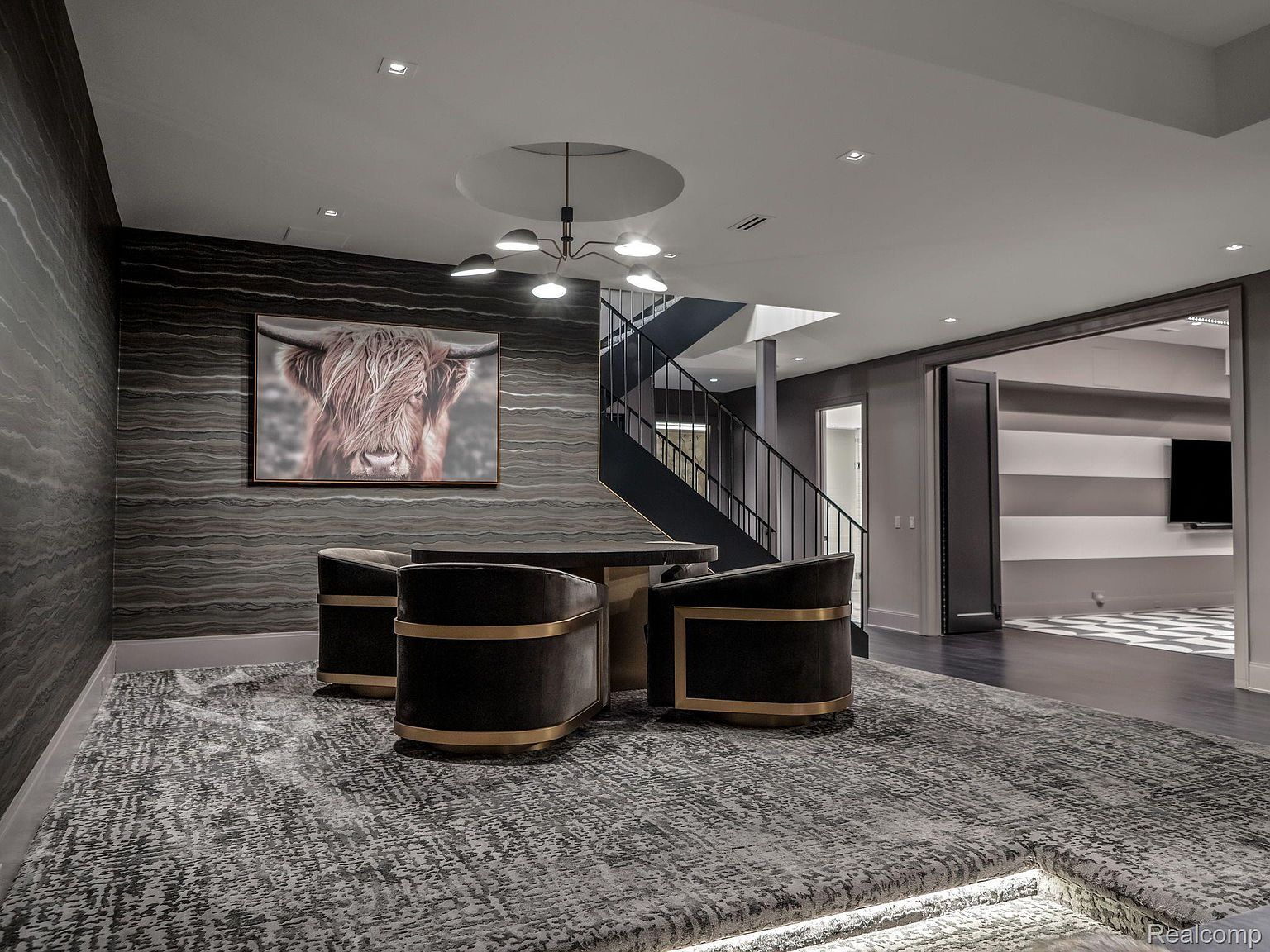 This is a stylish basement area featuring a seating arrangement with four chairs and a table, set upon a patterned rug on a raised platform. A large artwork of a highland cow adorns the wall, complemented by modern lighting fixtures and a staircase in the background. The space opens into another room with a black and white patterned floor, creating a sophisticated and inviting atmosphere.