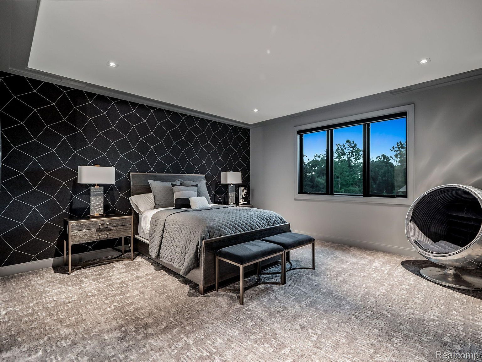 This is a primary bedroom featuring a modern design with a geometric patterned accent wall behind the bed. The room is furnished with a bed, two nightstands with lamps, and a seating area near a window offering natural light. The color palette is primarily gray and black, creating a sophisticated and calming atmosphere.