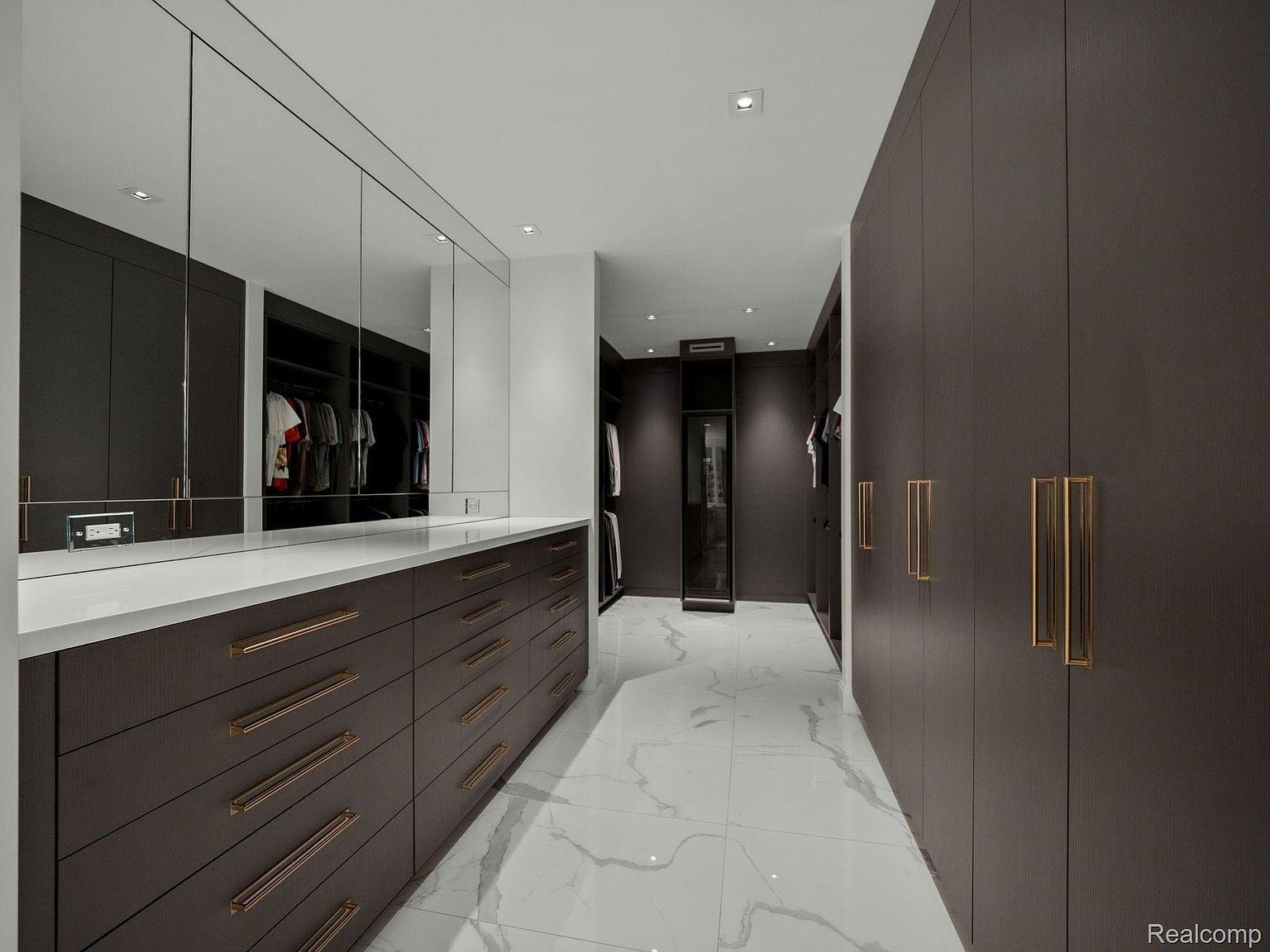 This is a well-organized walk-in closet featuring dark wood cabinetry with gold hardware and a white countertop. The closet includes drawers, shelving, and hanging space, with mirrors above the countertop area. The flooring is white marble with gray veining, contributing to a luxurious and modern aesthetic.