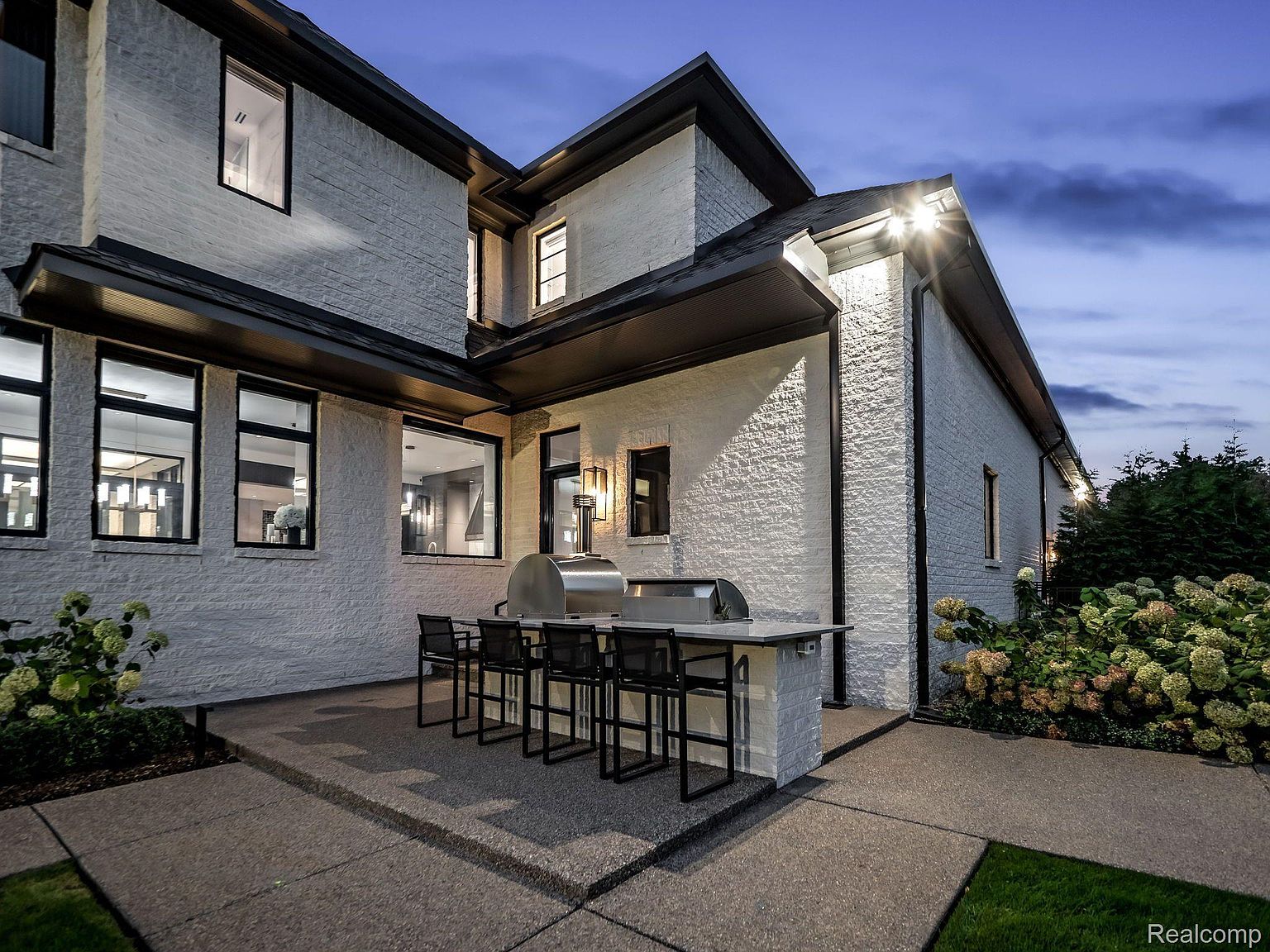 This image showcases the rear exterior of a modern home, featuring white brick walls, black-framed windows, and a dark roofline. An outdoor kitchen area with a built-in grill and bar seating is visible on a concrete patio. Lush landscaping adds a touch of nature to the scene, creating an inviting outdoor space.