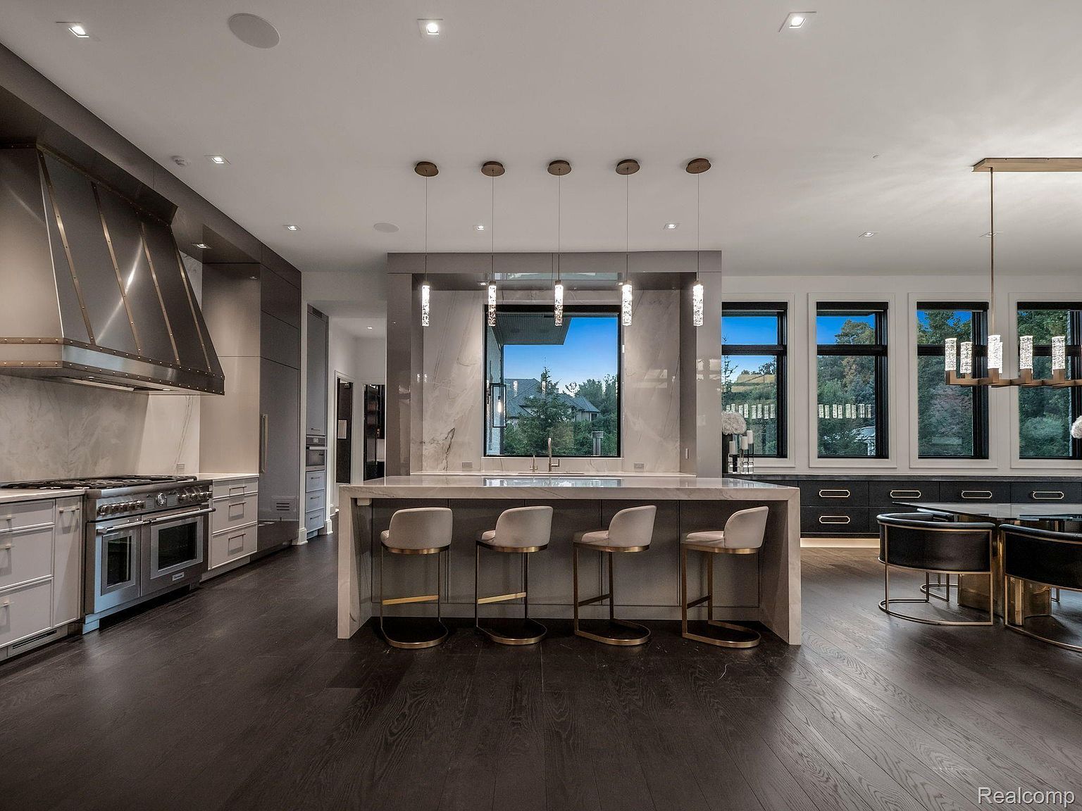 This is a wide shot of a modern, high-end kitchen featuring dark wood floors, a large island with seating, and stainless steel appliances. The kitchen is well-lit with recessed lighting and pendant lights over the island, and large windows provide natural light and views of the outdoors. The overall impression is one of luxury and sophistication.