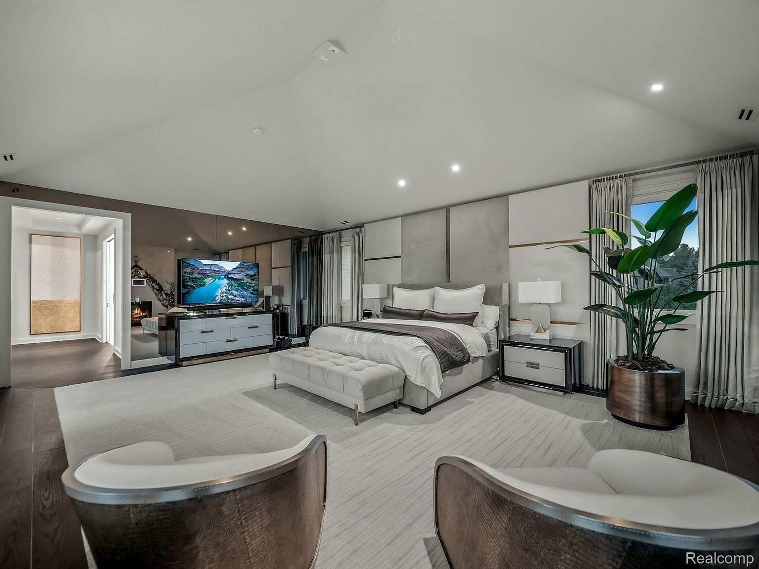 This is a luxurious primary bedroom featuring a large bed with neutral bedding, flanked by matching nightstands and lamps. A mirrored wall behind the TV creates a sense of spaciousness, while a seating area with two chairs and a bench offers a comfortable retreat. The room is well-lit with recessed lighting and natural light from the window, creating a serene and elegant atmosphere.