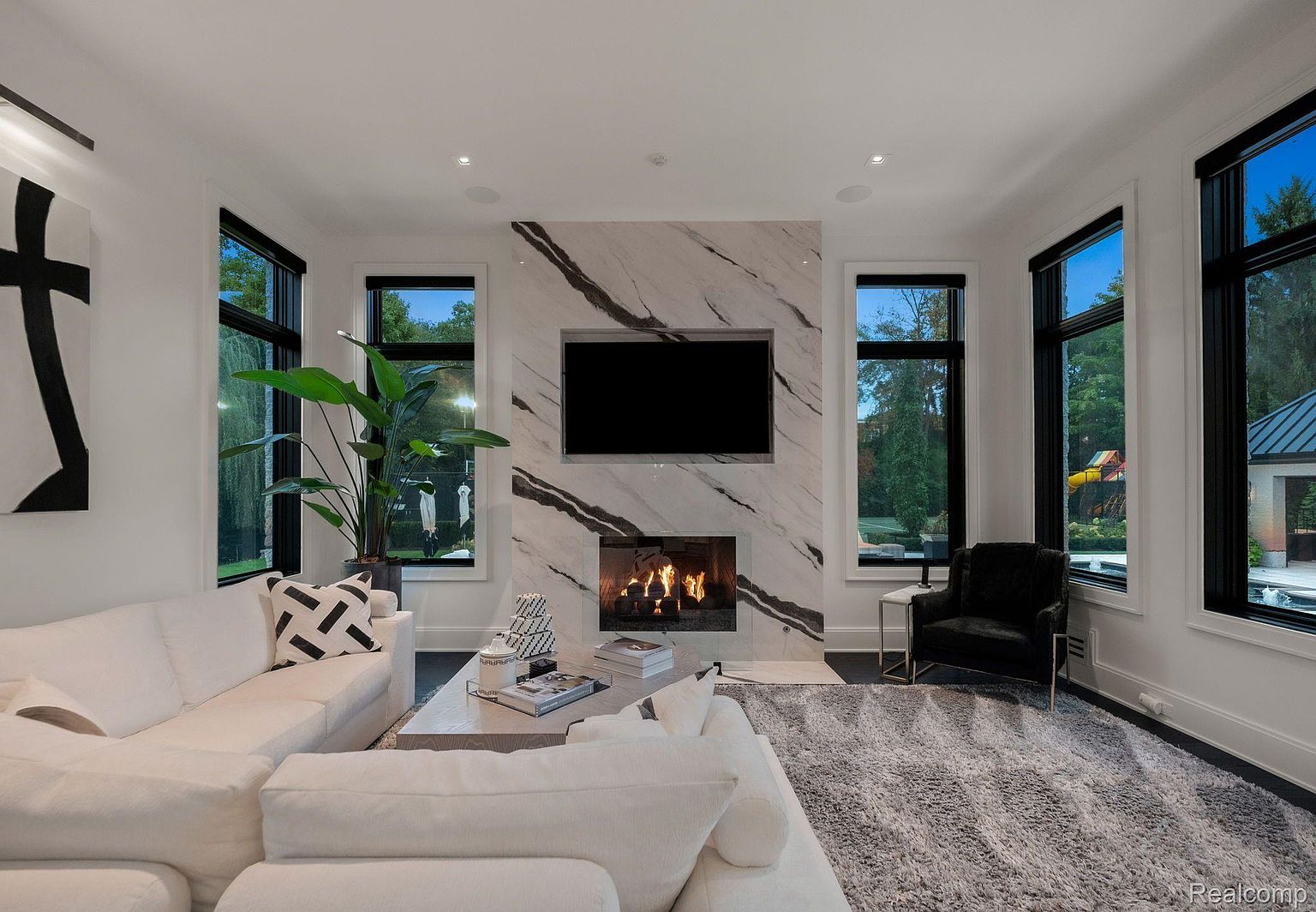 This is a bright and modern living room featuring a white sectional sofa, a marble fireplace with a built-in TV, and large windows that offer ample natural light. The room is decorated with contemporary art and accessories, creating a stylish and inviting atmosphere. A gray rug anchors the space, adding texture and warmth.