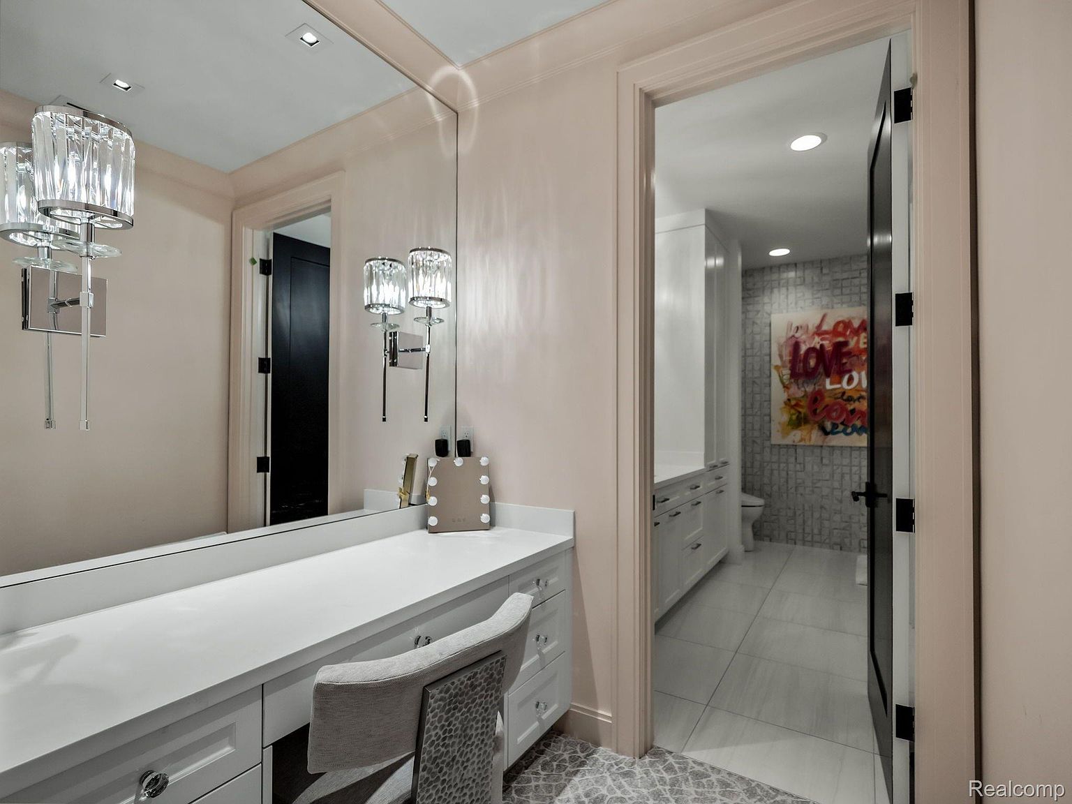 This is a primary bathroom featuring a vanity area with a large mirror, crystal sconces, and a white countertop with drawers. Through an open doorway, another section of the bathroom is visible, showcasing a white vanity, tiled walls, and a piece of art. The overall aesthetic is elegant and well-lit.