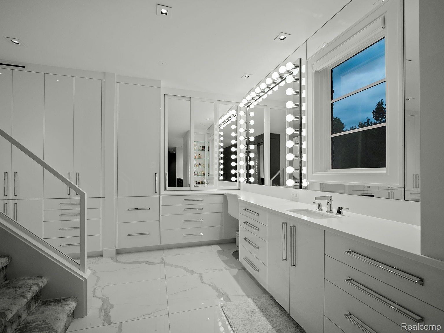 This is a luxurious primary bathroom featuring a large vanity with multiple mirrors and Hollywood-style lighting. The room is predominantly white, with sleek cabinetry, chrome hardware, and marble flooring. A window provides natural light, and the overall impression is clean, modern, and upscale.