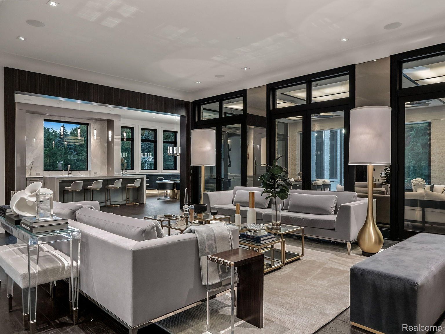 This is an interior shot of a luxurious living room, featuring two gray sofas, glass and gold coffee tables, and a gray ottoman. The room has large windows with black frames, allowing natural light to illuminate the space. The open-concept design connects the living room to a modern kitchen with bar seating, creating a seamless flow.