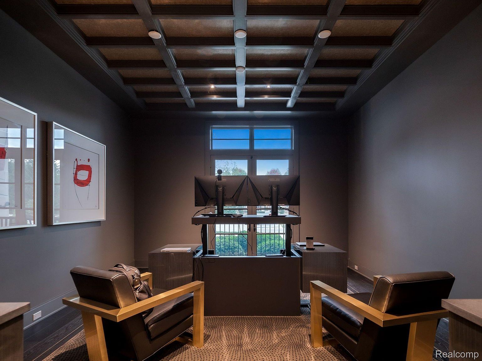 This is an interior shot of a modern office or study. The room features dark walls, a coffered ceiling with recessed lighting, and a large window providing natural light. A standing desk with dual monitors is positioned in front of the window, flanked by two leather chairs with wooden frames, creating a sophisticated and functional workspace.