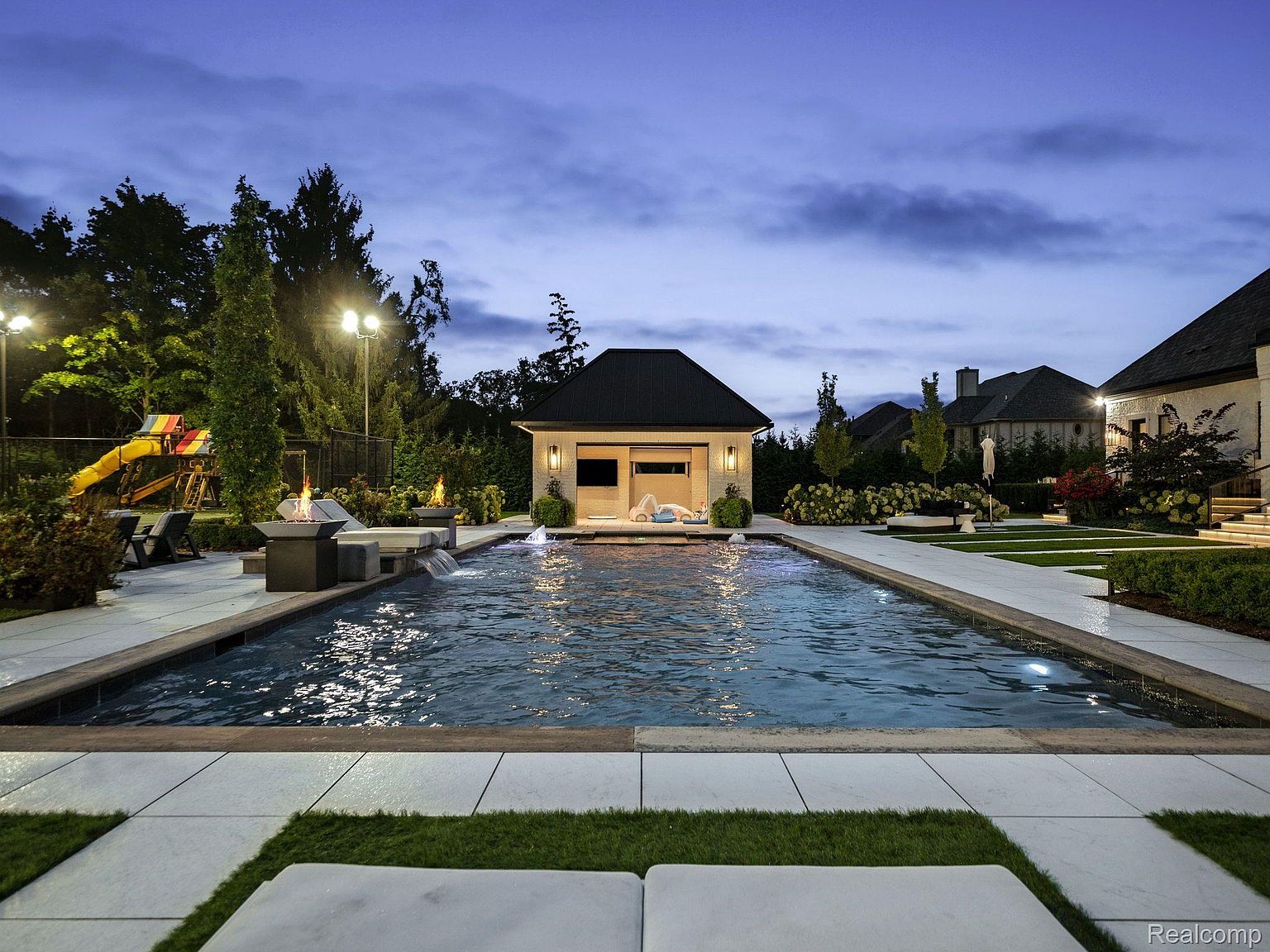 This image showcases a luxurious backyard pool area at dusk. The pool is surrounded by white tile and features a modern design with a small waterfall feature. A pool house sits at the far end, and fire pits add a touch of warmth and ambiance. The scene is framed by lush landscaping and elegant outdoor lighting, creating a serene and inviting atmosphere.