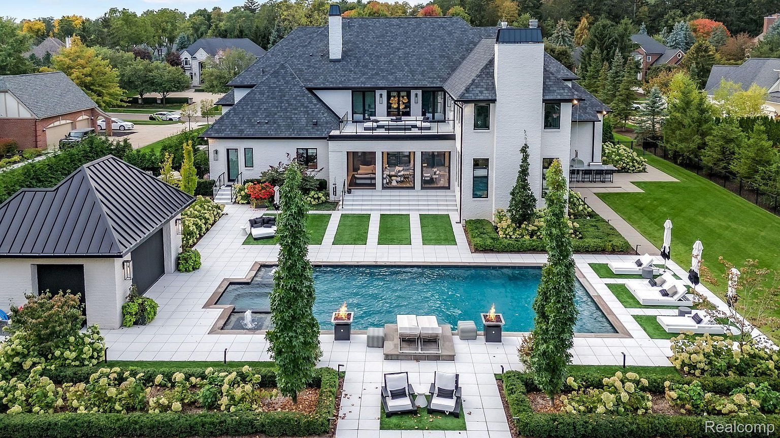 This is a rear view of a luxurious modern home, showcasing a pristine backyard with a large swimming pool, a patio area with lounge chairs, and meticulously manicured landscaping. The house features a white brick exterior, a dark roof, and a second-story balcony, creating an impression of elegance and sophistication. The perspective is from an elevated position, providing a comprehensive view of the property's outdoor living spaces.