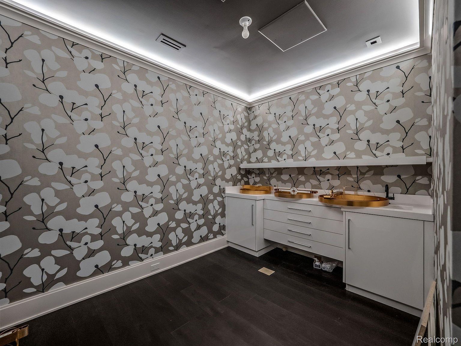 This is a well-lit bathroom featuring a floral wallpaper design, dark wood flooring, and a double vanity with gold-colored sinks. The vanity has white cabinets and drawers, and a shelf is mounted above it. The room has a modern and stylish aesthetic.
