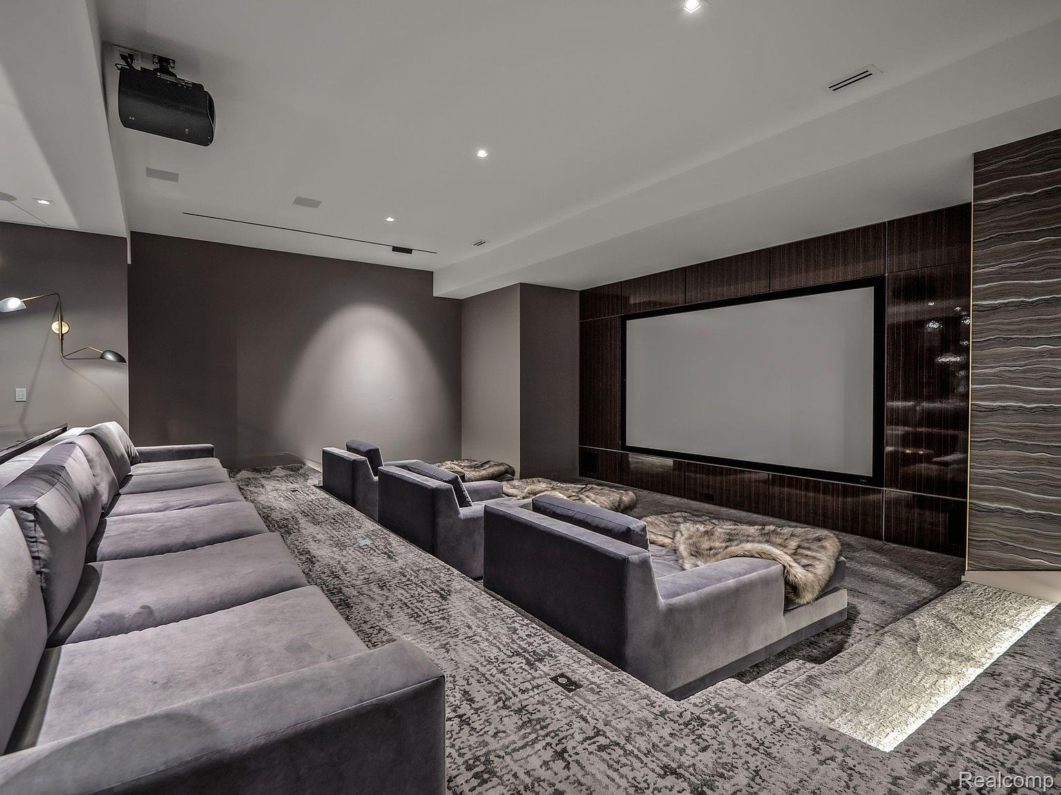 This is a luxurious home theater room featuring tiered seating with plush, gray upholstered sofas and armchairs. A large screen is mounted on a dark wood-paneled wall, complemented by textured wall accents. The room is carpeted with a patterned gray rug, and the overall ambiance is sophisticated and comfortable.