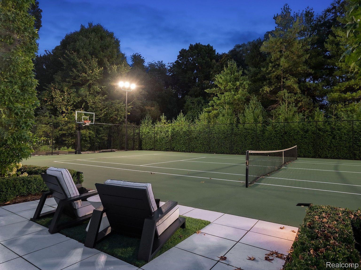 This image showcases a well-maintained tennis and basketball court at dusk, illuminated by overhead lighting. Two modern lounge chairs sit on a paved patio adjacent to the court, offering a relaxing space for spectators. The surrounding landscape features mature trees and manicured hedges, creating a private and luxurious outdoor recreational area.