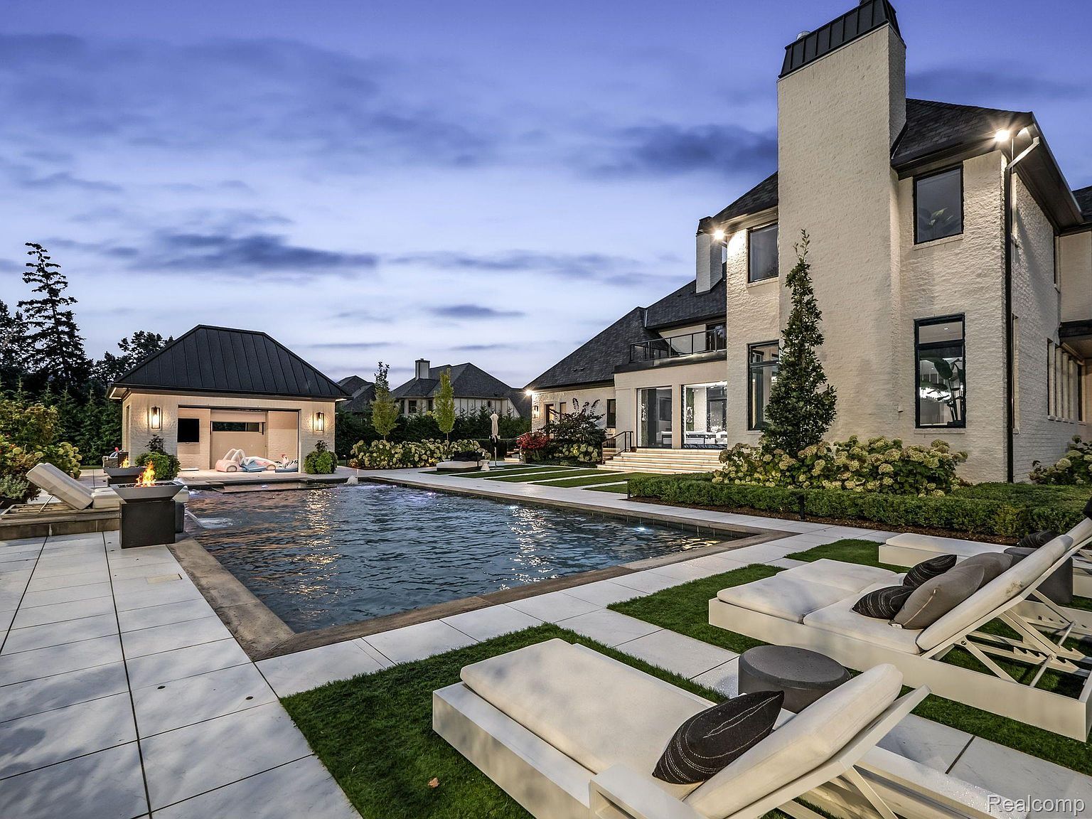 This image showcases a luxurious backyard featuring a modern pool and spa area. The pool is surrounded by white tile and lush green grass, complemented by stylish lounge chairs with decorative pillows. A pool house with a dark roof sits adjacent to the pool, and the main house, a large white brick structure with a prominent chimney, is visible in the background, creating an inviting and upscale outdoor living space.