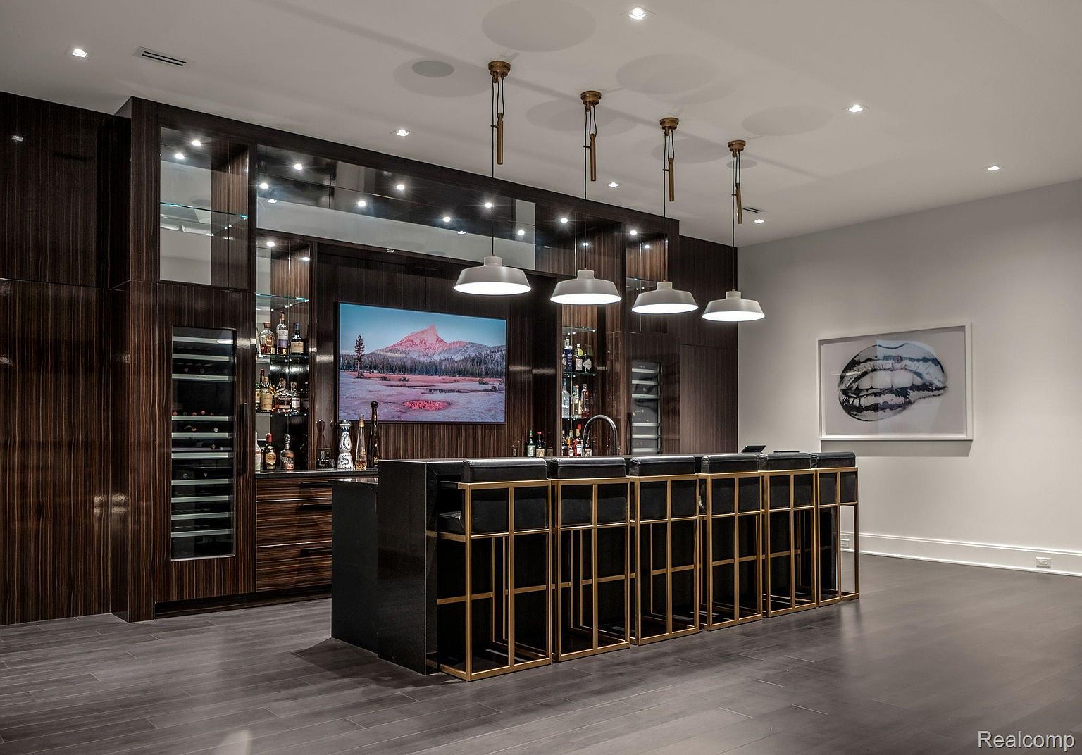 This interior shot showcases a sophisticated wine cellar and bar area. The dark wood cabinetry, mirrored accents, and modern pendant lighting create a luxurious ambiance. A sleek bar with black and gold stools stands in the foreground, complemented by a large television displaying a scenic landscape.