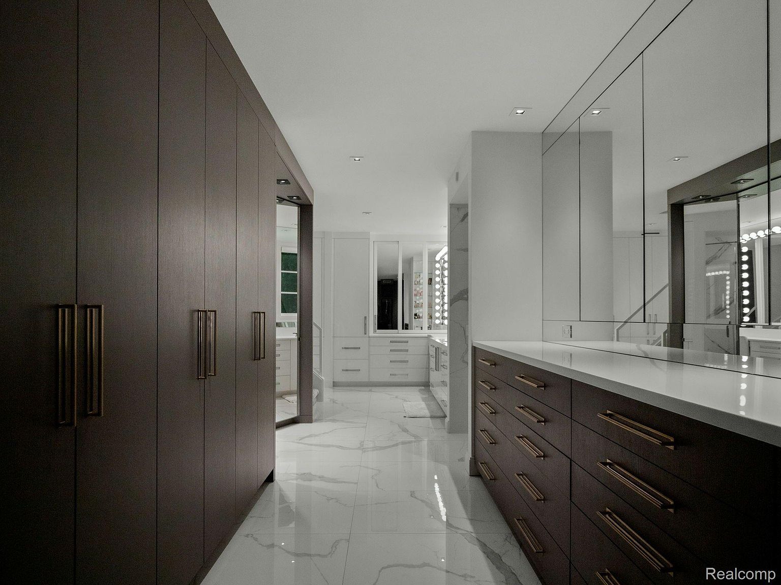 This is a luxurious walk-in closet featuring dark wood cabinetry with gold hardware and a white marble floor. The closet includes ample storage space and a vanity area with mirrors and lighting. The overall impression is one of high-end design and organization.