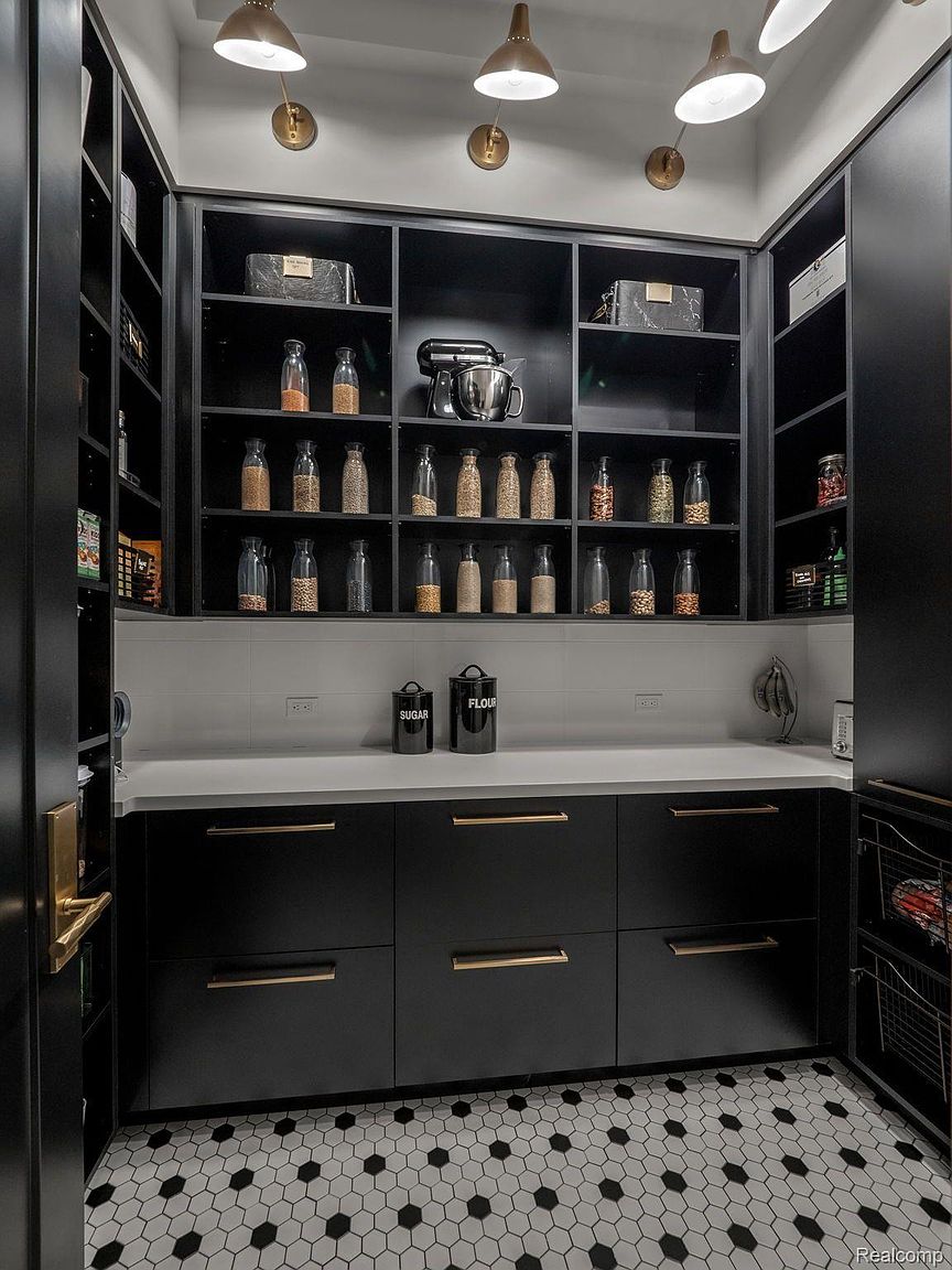 This is a well-organized pantry featuring black cabinetry with gold hardware and a white countertop. The shelves are stocked with various jars and containers, and a stand mixer is visible. The floor is tiled with a black and white hexagonal pattern, and the overall impression is one of modern elegance and functionality.