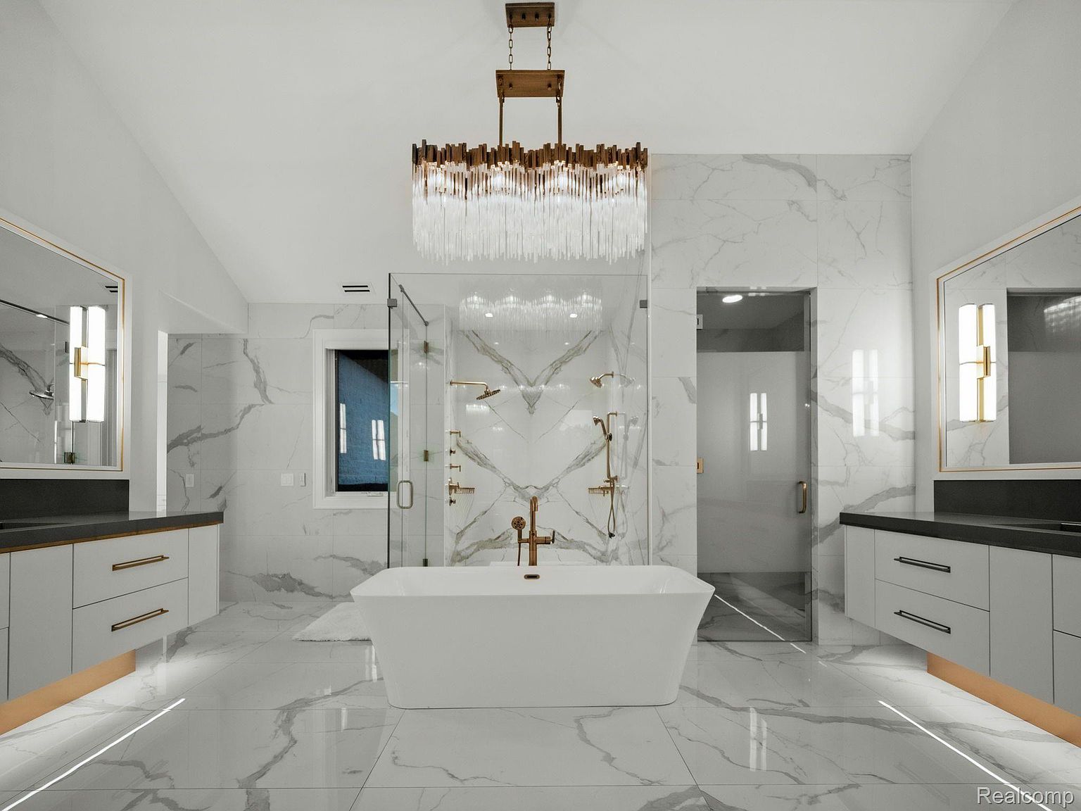 This is a luxurious primary bathroom featuring marble-like tile flooring and walls, a freestanding white bathtub, and a modern chandelier. The bathroom includes dual vanities with dark countertops and gold accents, as well as a glass-enclosed shower and a separate toilet room. The overall impression is one of elegance and spa-like tranquility.