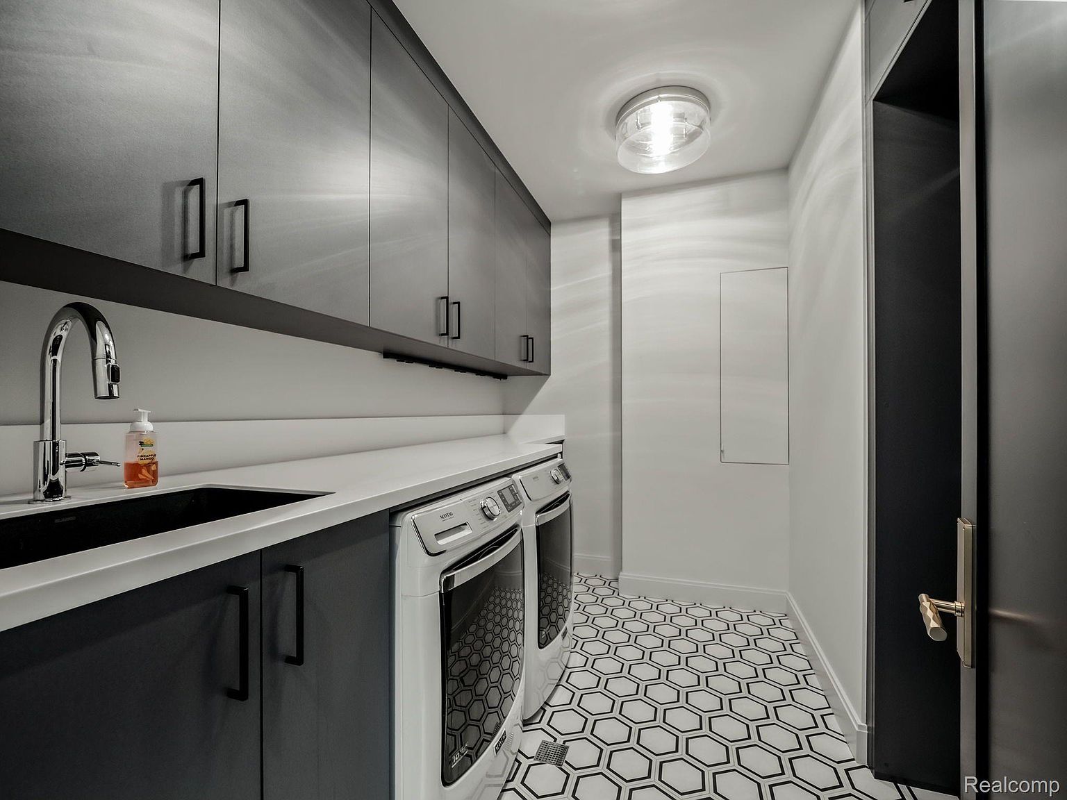 This is a modern laundry room featuring sleek, dark gray cabinetry with minimalist hardware and a white countertop. A stainless steel faucet complements the dark sink, and a white front-loading washer and dryer are neatly installed. The black and white hexagonal tile flooring adds a stylish touch to the space, which is well-lit by a flush-mount ceiling light.