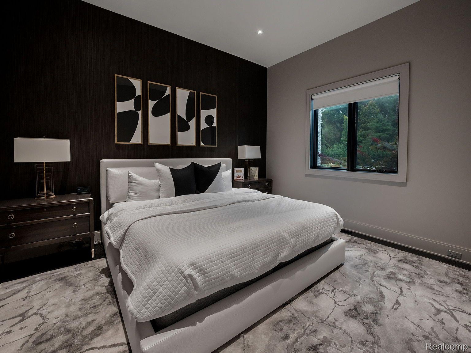 This is a primary bedroom featuring a king-size bed with white bedding and black accent pillows, set against a dark accent wall adorned with modern art. The room includes matching nightstands with lamps, and a window providing natural light. The flooring is a unique marble pattern, adding a touch of luxury to the space.