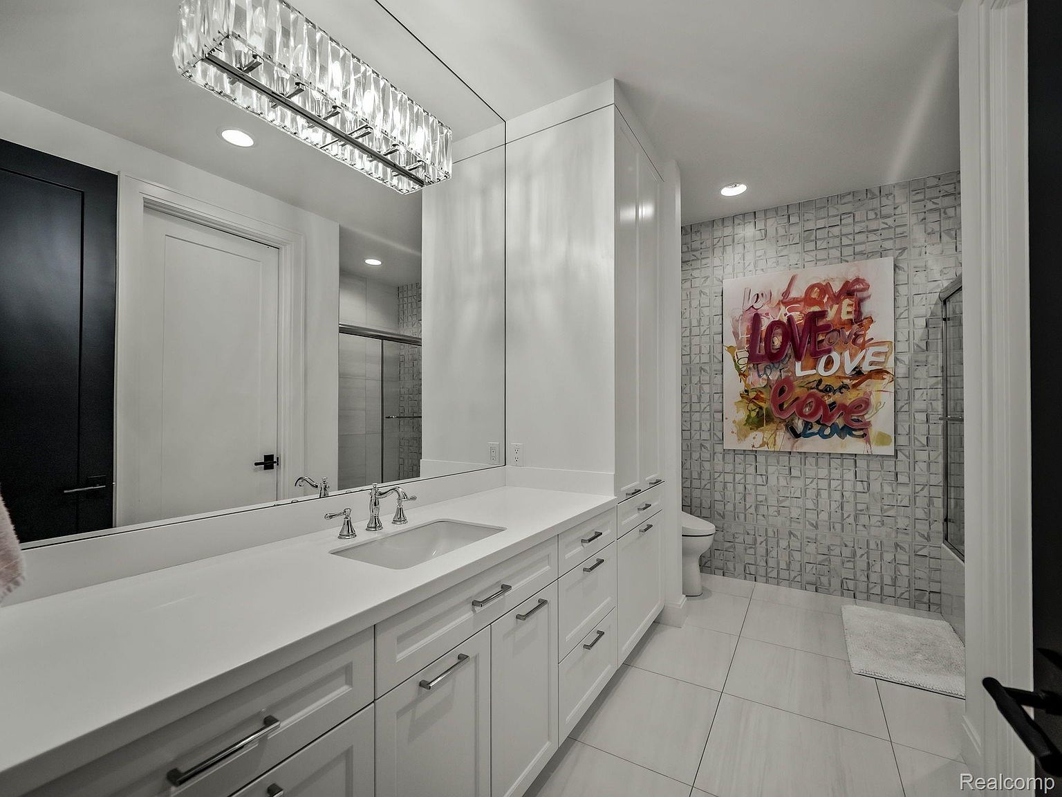 This is a well-lit primary bathroom featuring a long white vanity with ample storage, a large mirror with a modern chandelier, and a separate shower area. The walls are decorated with a textured tile and a colorful 'LOVE' artwork, adding a touch of personality to the space. The overall impression is clean, modern, and stylish.
