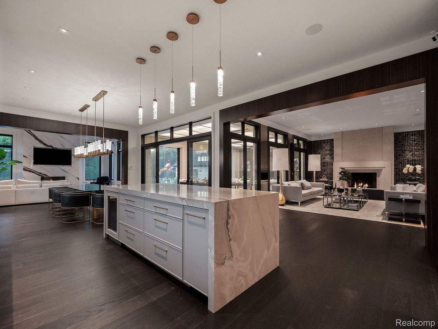 This is a wide-angle shot of a luxurious kitchen and living area. The kitchen features a large island with marble countertops and modern cabinetry, illuminated by pendant lighting. The living area includes a fireplace and comfortable seating, creating an open and inviting space with dark wood floors throughout.