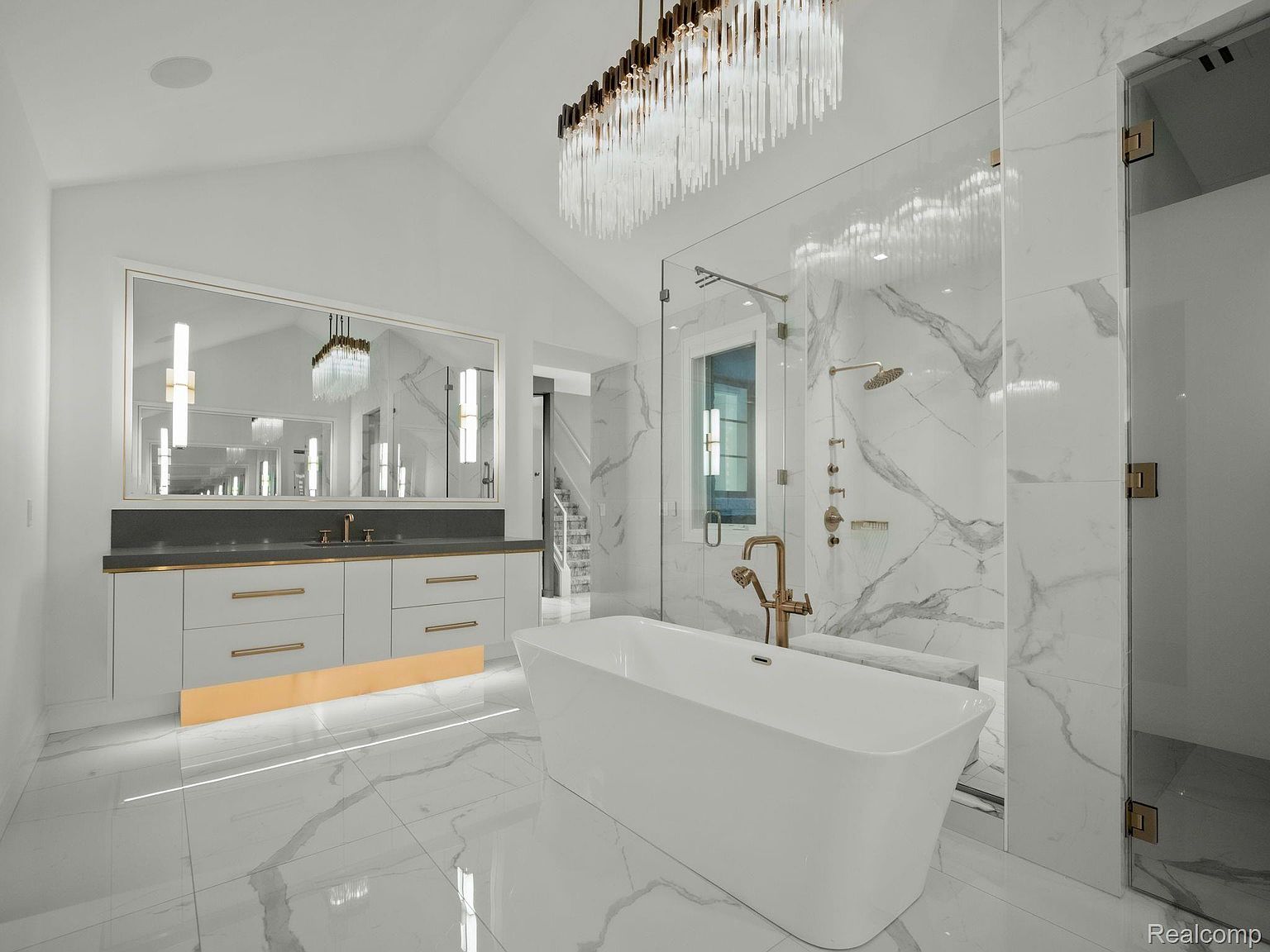 This is a luxurious primary bathroom featuring a freestanding white bathtub as a focal point. The walls and floors are covered in marble-patterned tiles, and a glass-enclosed shower is visible in the background. A modern vanity with gold accents and a large mirror complete the elegant and spa-like atmosphere.