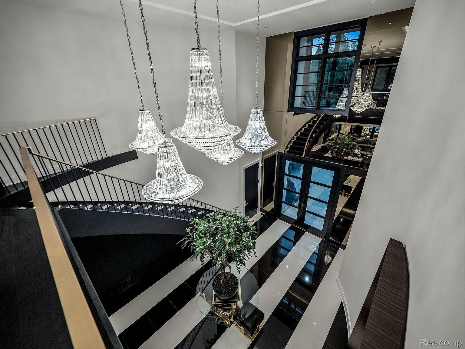 This is an interior shot of a grand foyer featuring a striking black and white striped tile floor, a curved staircase with black railings, and several ornate crystal chandeliers hanging from the ceiling. A large window and mirrored wall reflect the scene, enhancing the sense of space and luxury. The overall impression is one of modern elegance and sophisticated design.