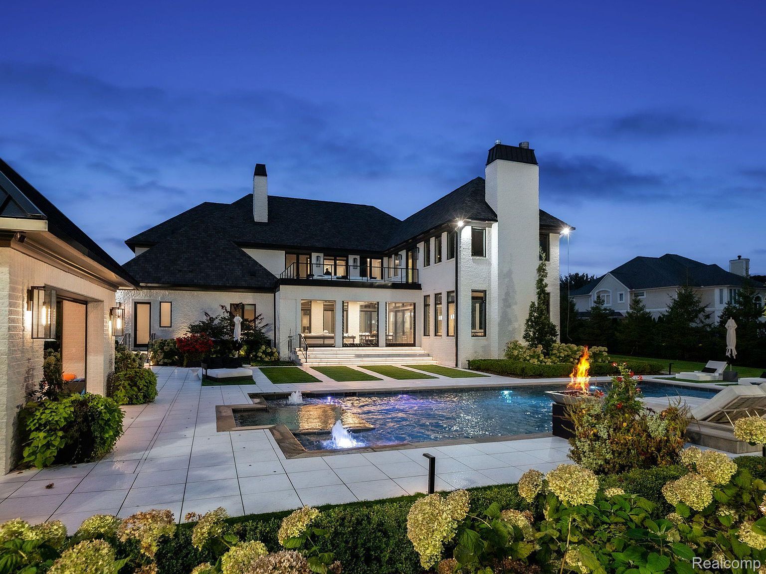 This image showcases the rear exterior of a luxurious home, highlighting a stunning pool and spa area. The pool features illuminated water fountains, and a fire pit adds warmth and ambiance. The surrounding patio is spacious and well-manicured, with lush landscaping and elegant outdoor furniture, creating an inviting and upscale outdoor living space.