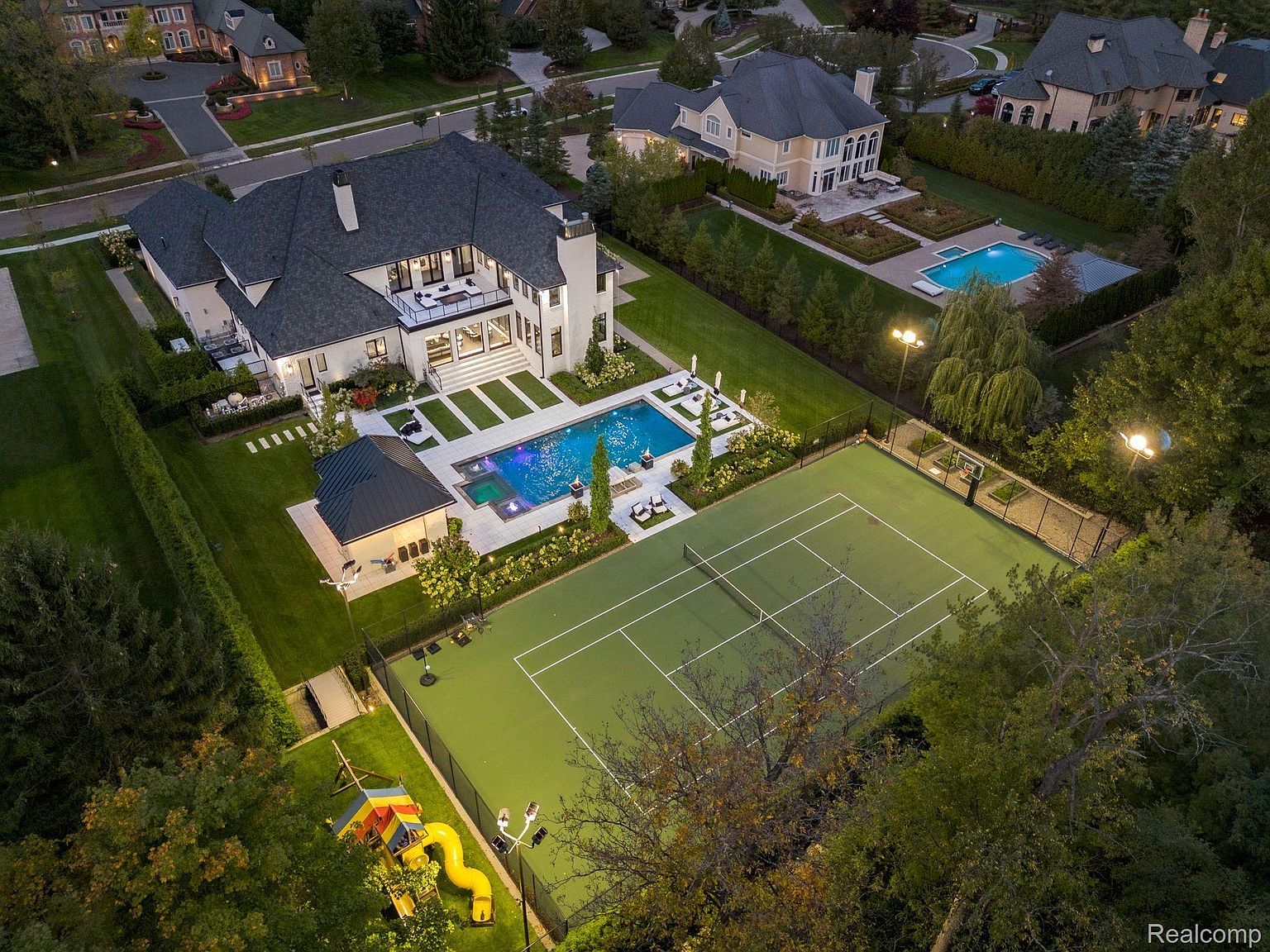 This aerial view showcases a luxurious estate featuring a large, modern home with a dark roof and white exterior. The property includes a swimming pool with a spa, a tennis court, a pool house, and a meticulously landscaped yard. The perspective highlights the scale and high-end amenities of this impressive residence.
