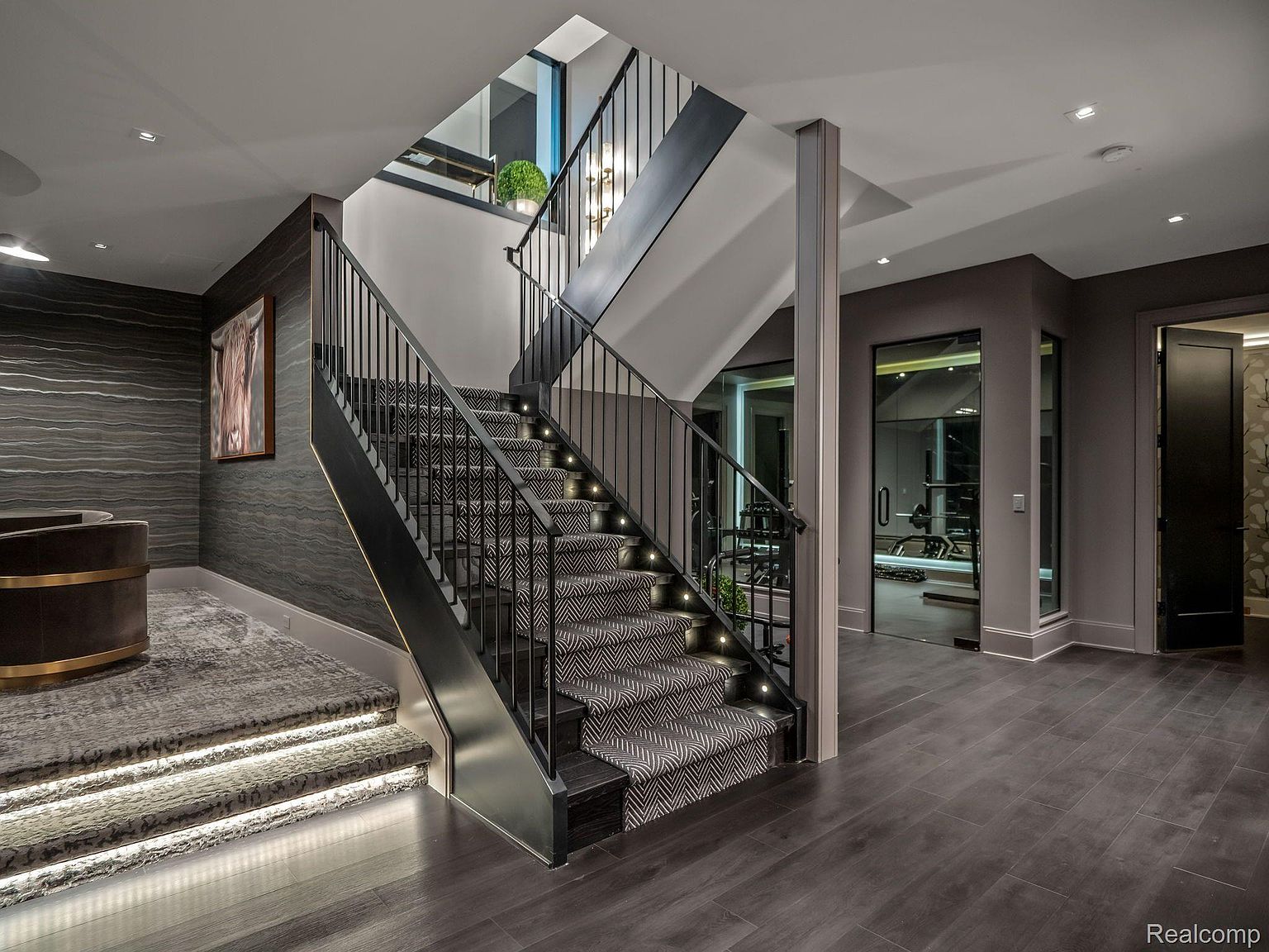 This interior shot showcases a modern hallway and staircase. The staircase features a patterned carpet runner, integrated step lighting, and a sleek black railing. The hallway leads to a home gym visible through a glass door, and the overall aesthetic is sophisticated and well-lit, creating an inviting and luxurious atmosphere.