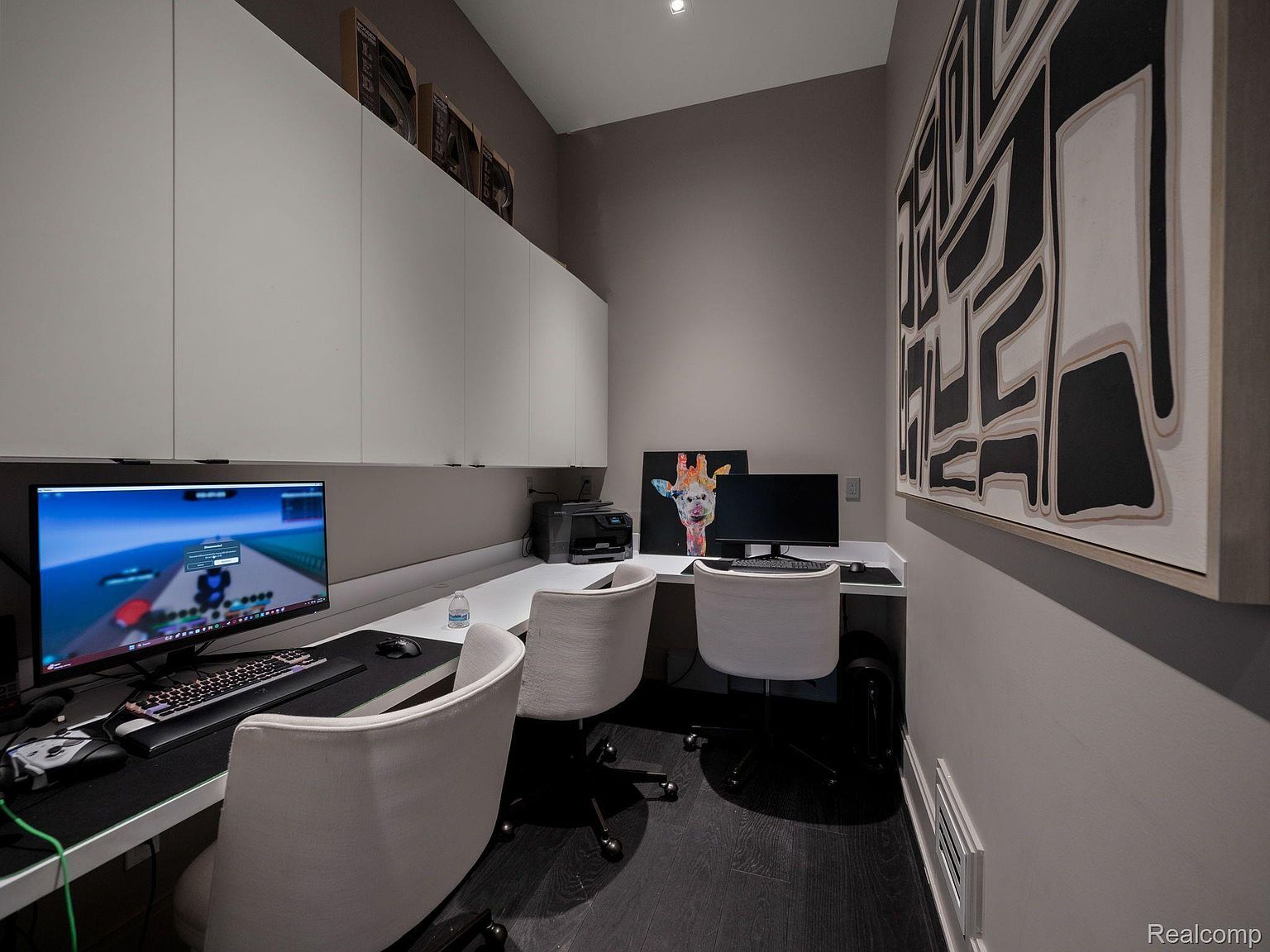 This is an interior shot of a modern office space featuring a built-in desk with two workstations. The room is decorated with a large abstract art piece and a colorful giraffe painting. The space is well-lit and organized, suggesting a productive and comfortable work environment.