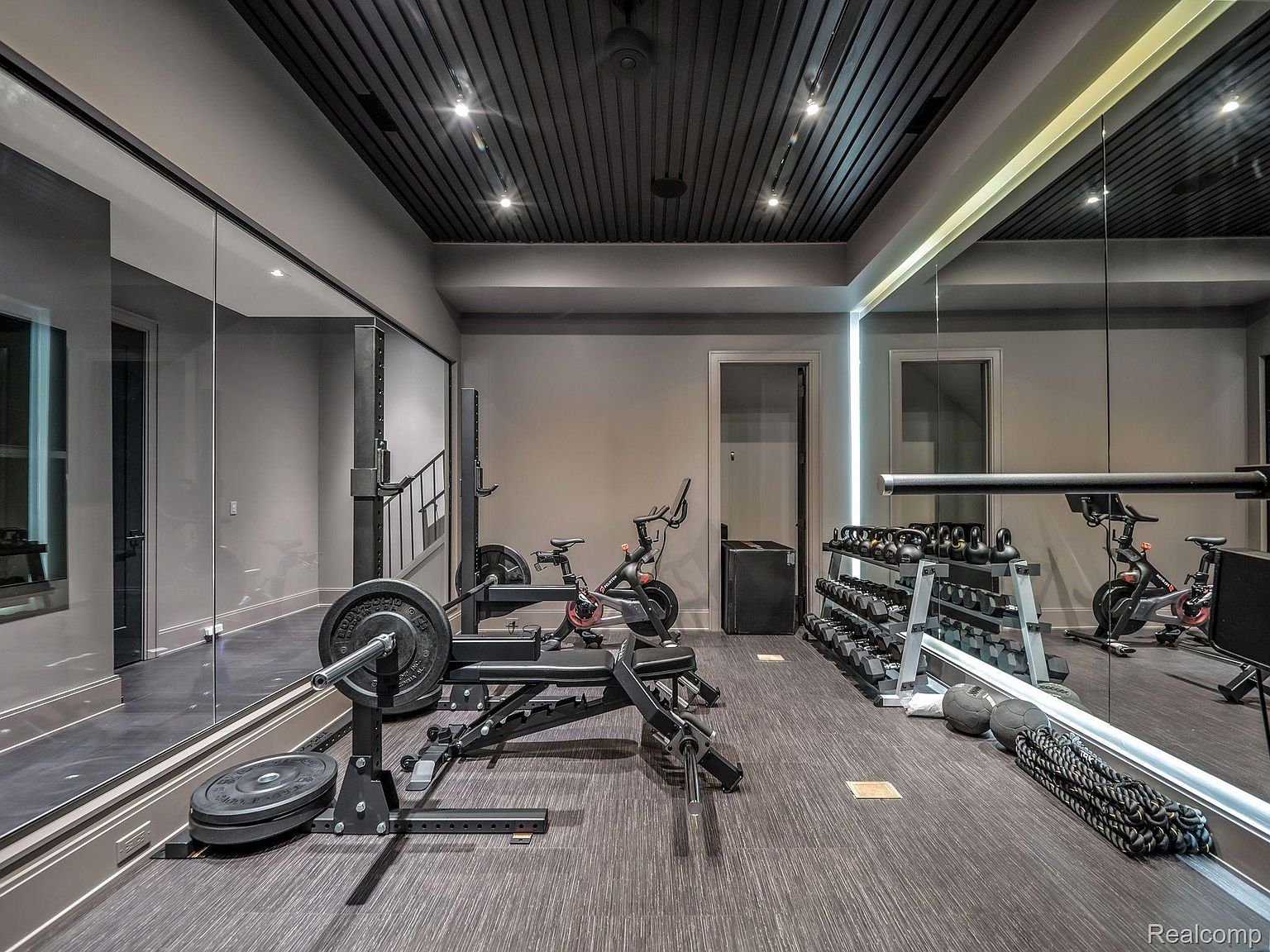 This is a well-equipped home gym featuring a weight bench with barbell, dumbbells, kettlebells, and exercise bikes. A large mirror enhances the space, and the dark ceiling with recessed lighting creates a modern aesthetic. The room is designed for a comprehensive workout experience.