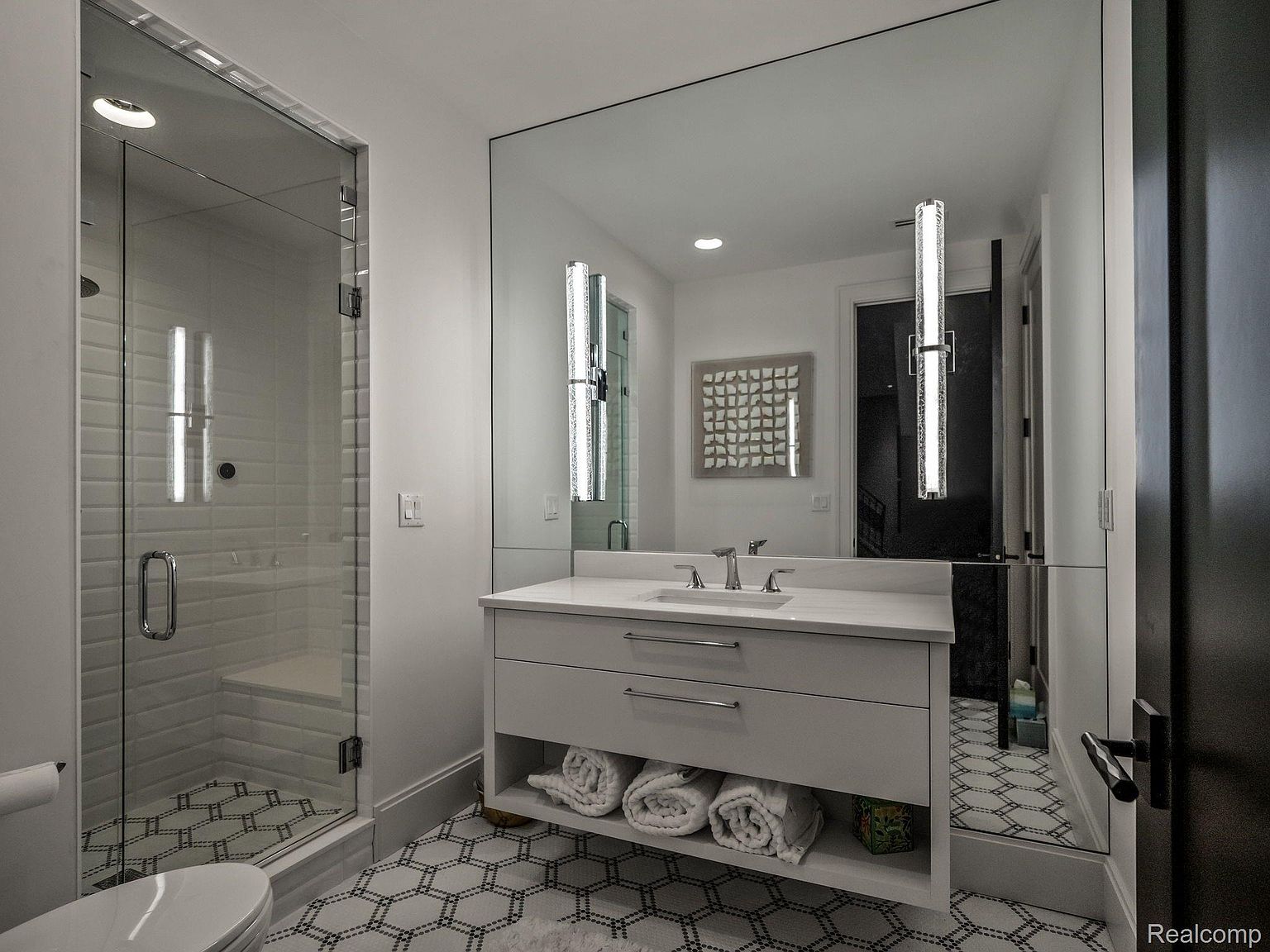 This is a well-lit bathroom featuring a glass-enclosed shower with white subway tile and a built-in bench. The vanity has a white countertop, gray cabinetry with drawers, and an open shelf for towel storage. The floor is tiled with a geometric pattern, and a large mirror reflects the space, enhancing the sense of openness.