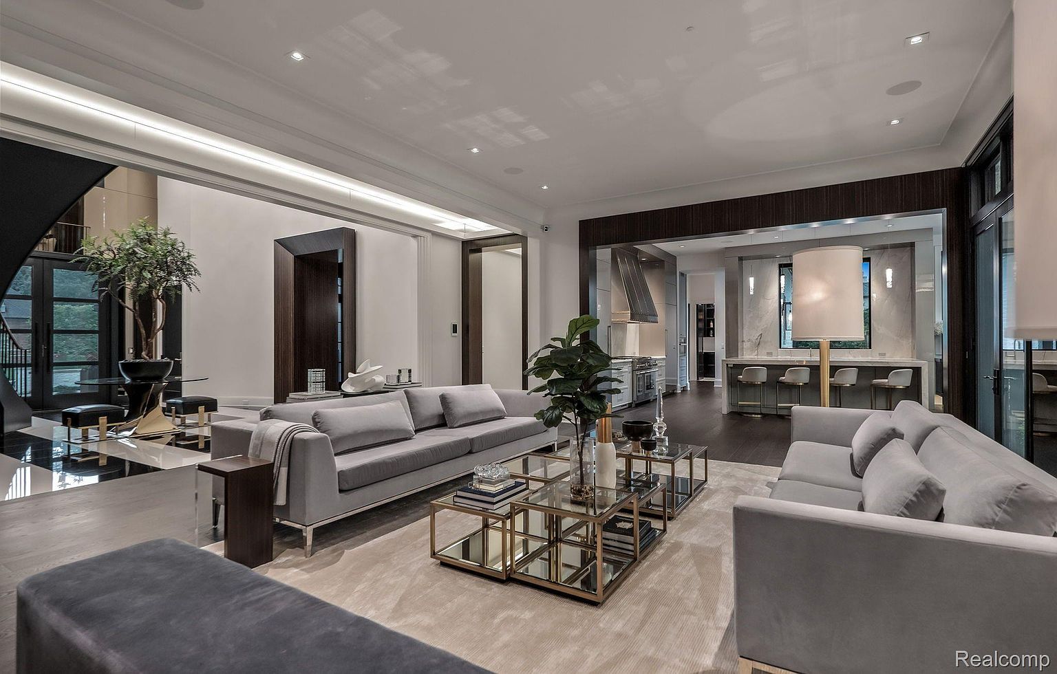 This is a spacious and luxurious living room featuring two gray sofas, a glass and gold coffee table, and a neutral-toned rug. The room is open to the kitchen area, creating a seamless flow. The design is modern and sophisticated, with high-end finishes and ample natural light.
