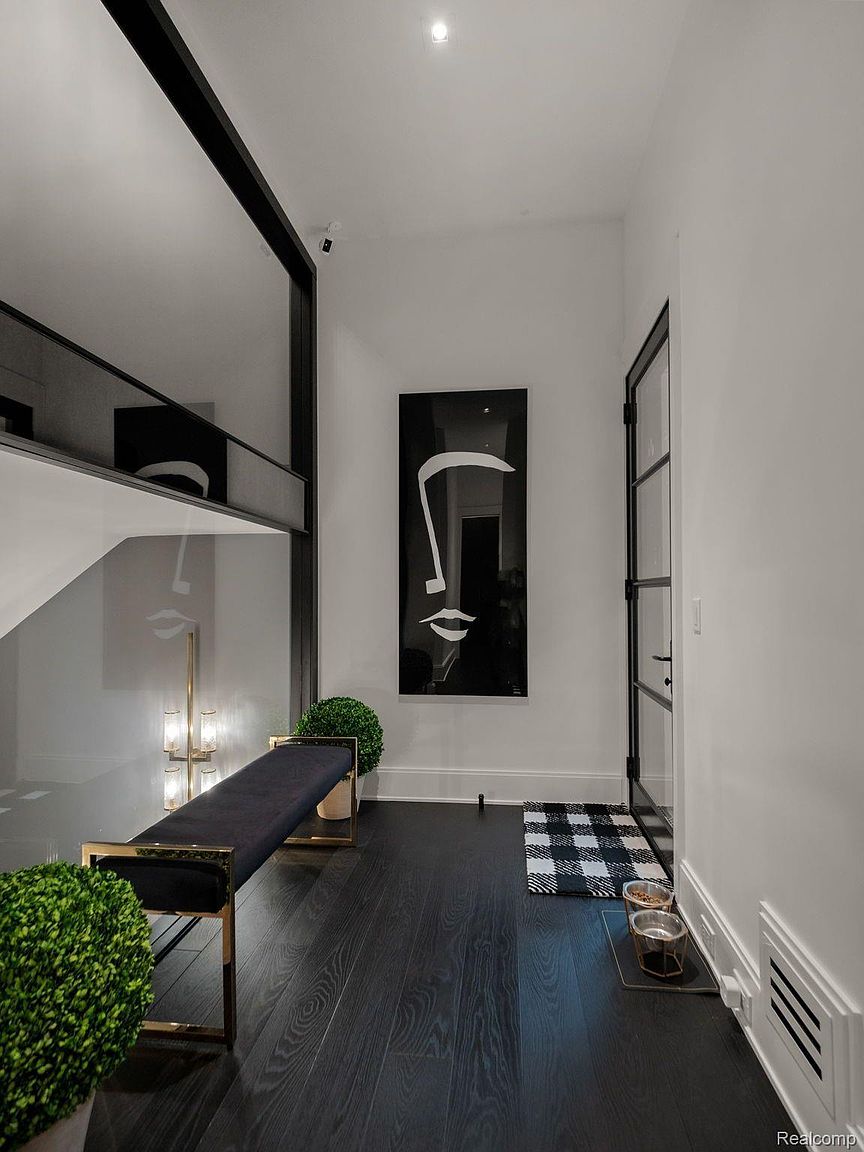 This is an interior shot of a modern hallway featuring dark hardwood floors and white walls. A black and gold bench sits against a mirrored wall, reflecting the staircase. A black and white art piece hangs on the wall, and a black-framed glass door is visible at the end of the hallway, creating a sleek and sophisticated aesthetic.