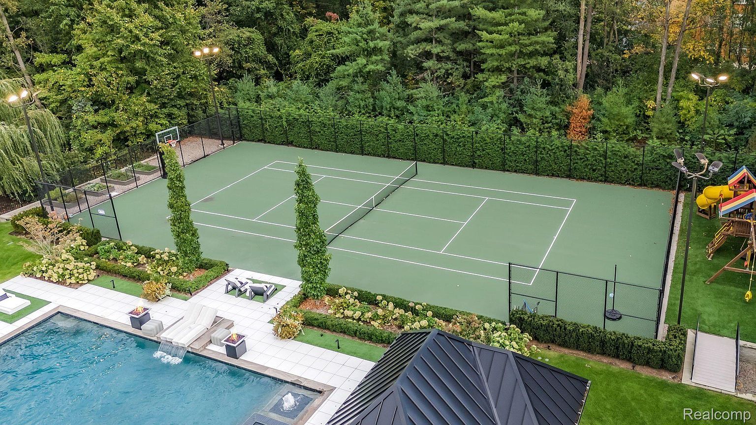 This aerial shot showcases a luxurious backyard featuring a tennis court, swimming pool, and meticulously landscaped gardens. The tennis court is surrounded by a black fence and lush greenery, while the pool area includes lounge chairs and a waterfall feature. The overall impression is one of high-end outdoor living and recreation.