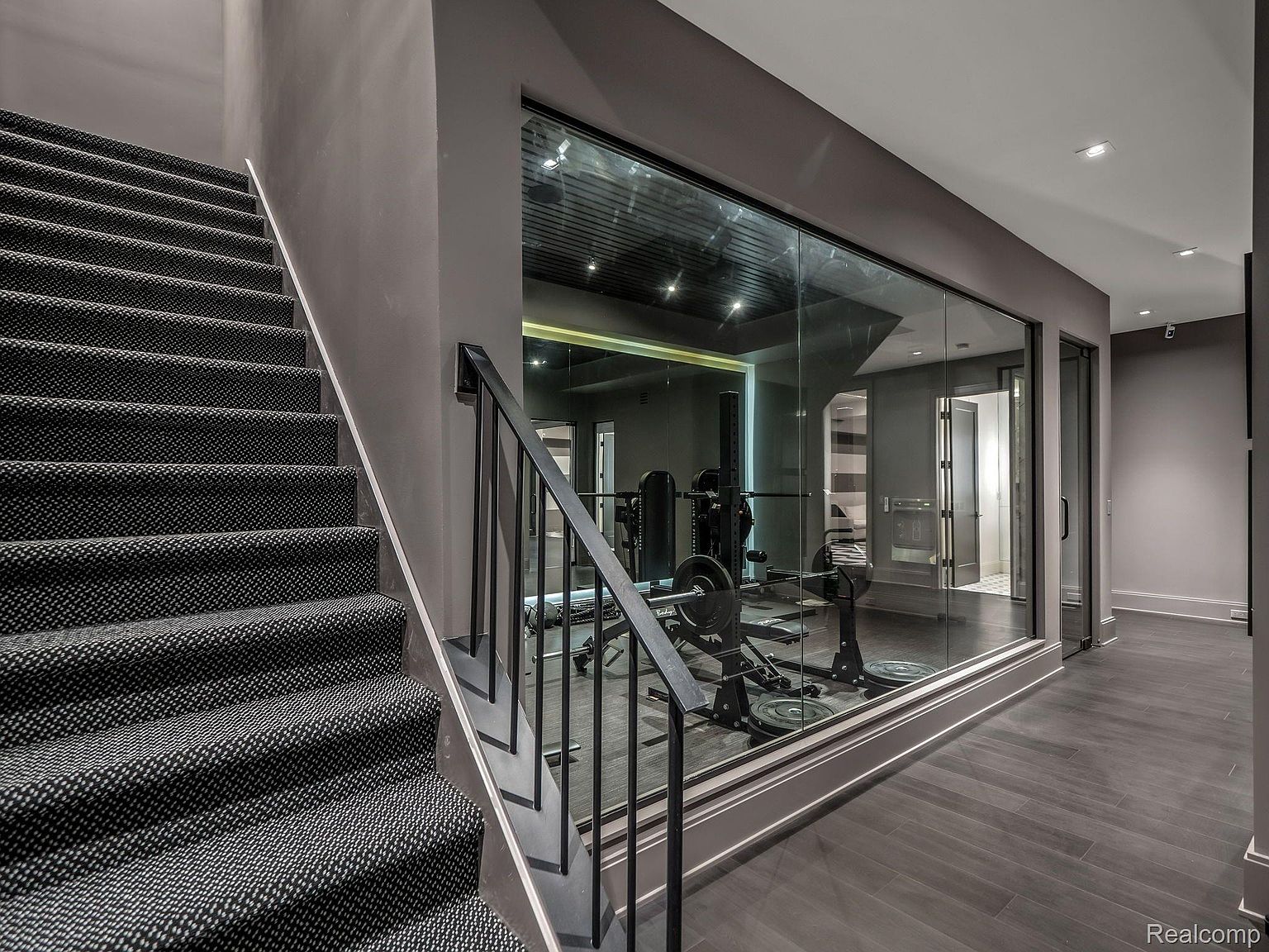 This image showcases a modern home gym viewed through a large glass partition, adjacent to a staircase. The gym features weightlifting equipment and a sleek, minimalist design with dark gray walls and flooring. The glass wall provides a clear view into the gym from the hallway, creating an open and inviting atmosphere.